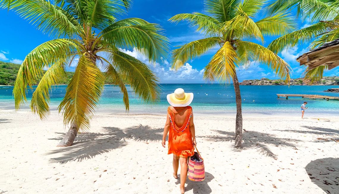 Woman carrying a tote bag and walking toward a beautiful turquoise ocean on a white sand beach with palm trees