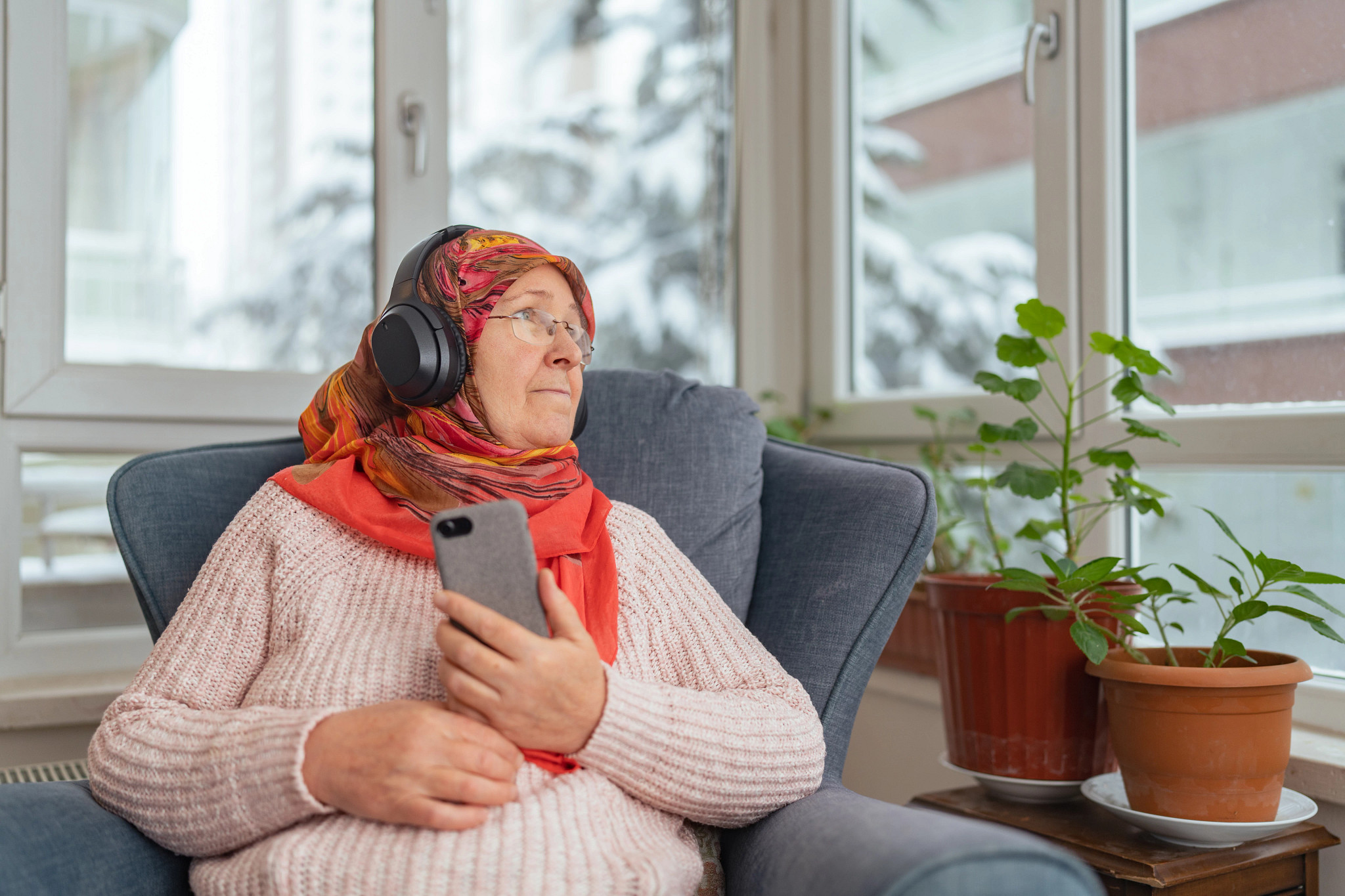 A muslim woman is sitting on sofa and relaxing and enjoying listening to music or podcast in the living room at home while it is snowing and cold outside.