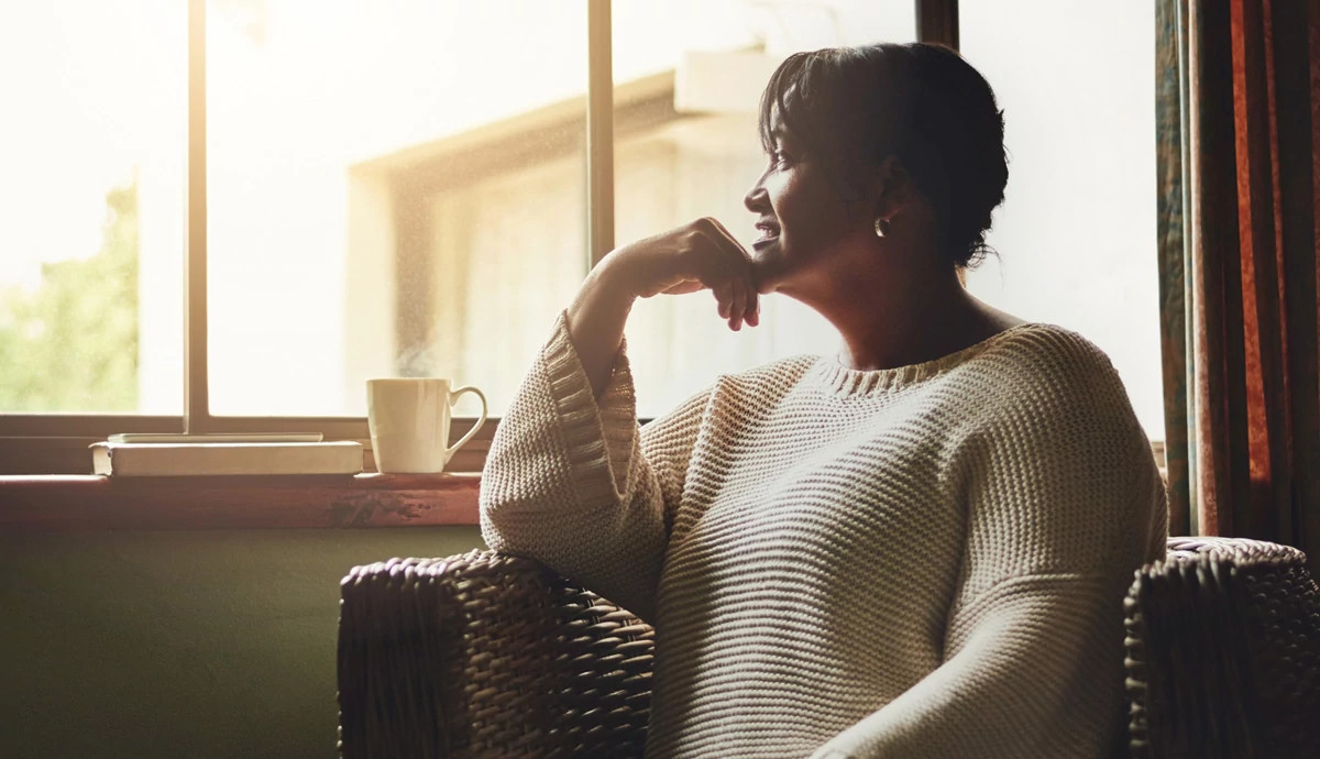 A photograph of a woman sitting in her living room and looking out the window.