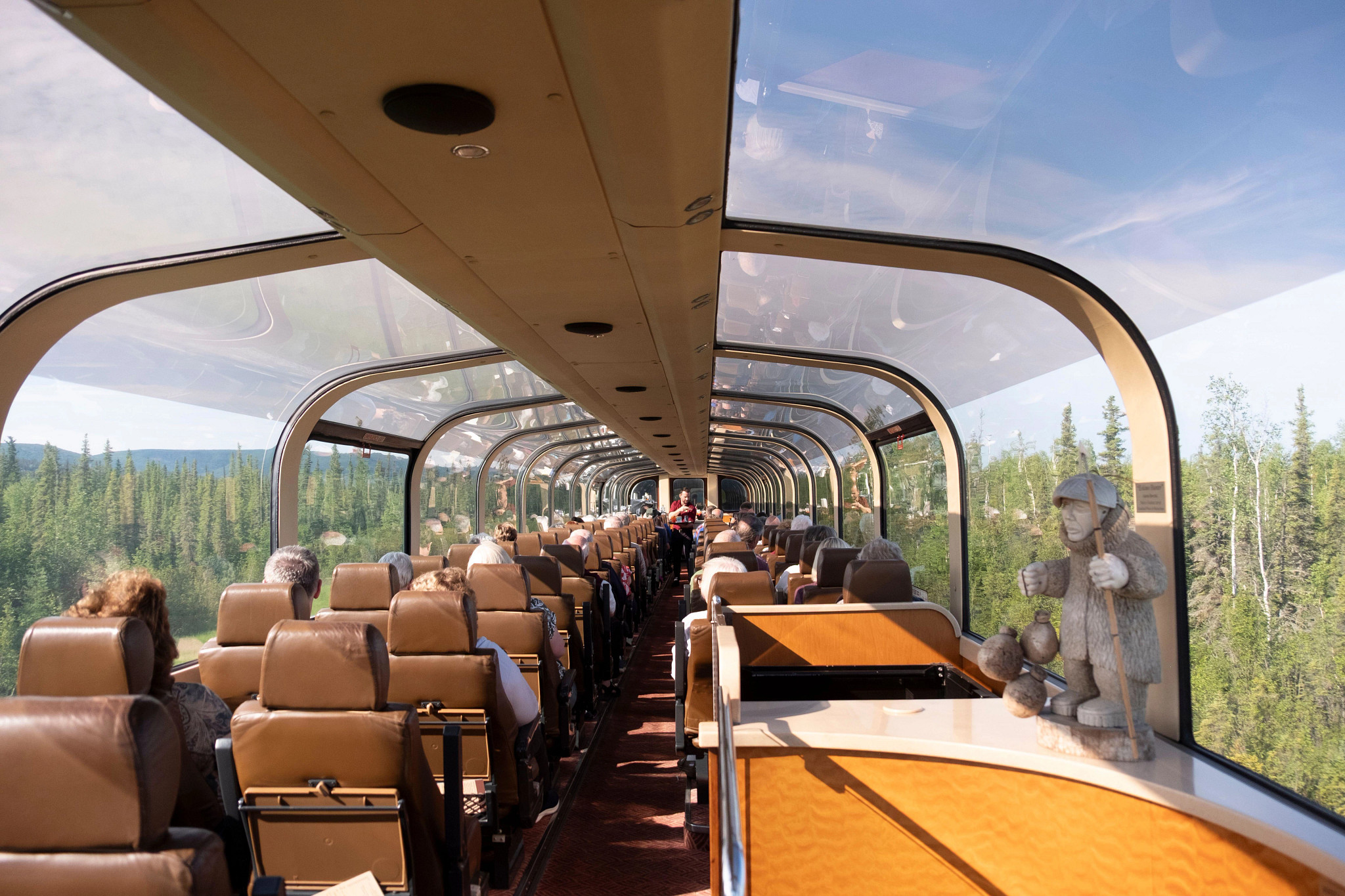 Passengers seated inside a panoramic glass-domed train observation car surrounded by forest scenery