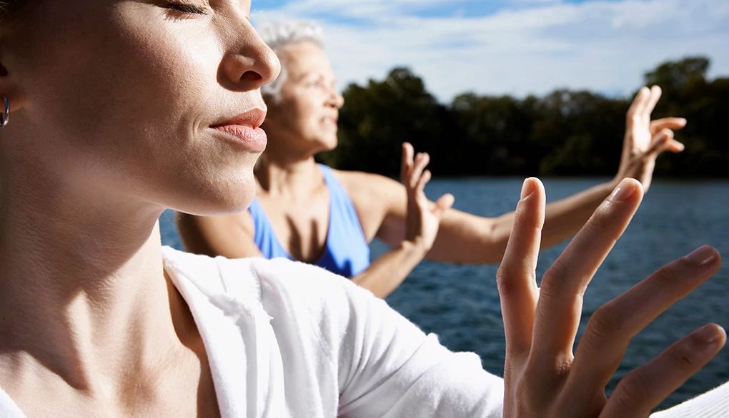 Tai chi may help support brain health A close-up view of two women doing tai chi by a lake