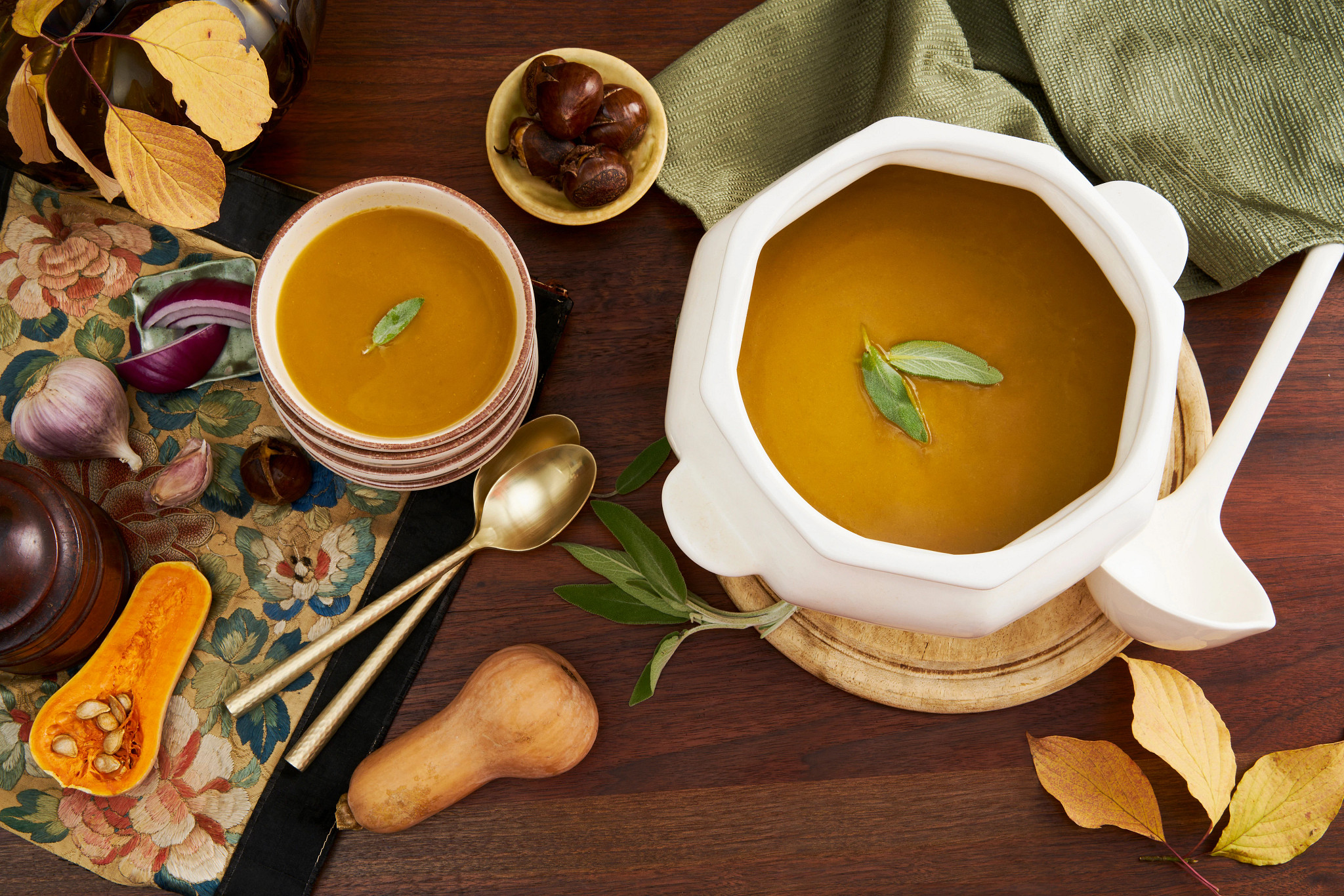 Golden holiday soup in a white tureen with sage garnish surrounded by autumn leaves and butternut squash