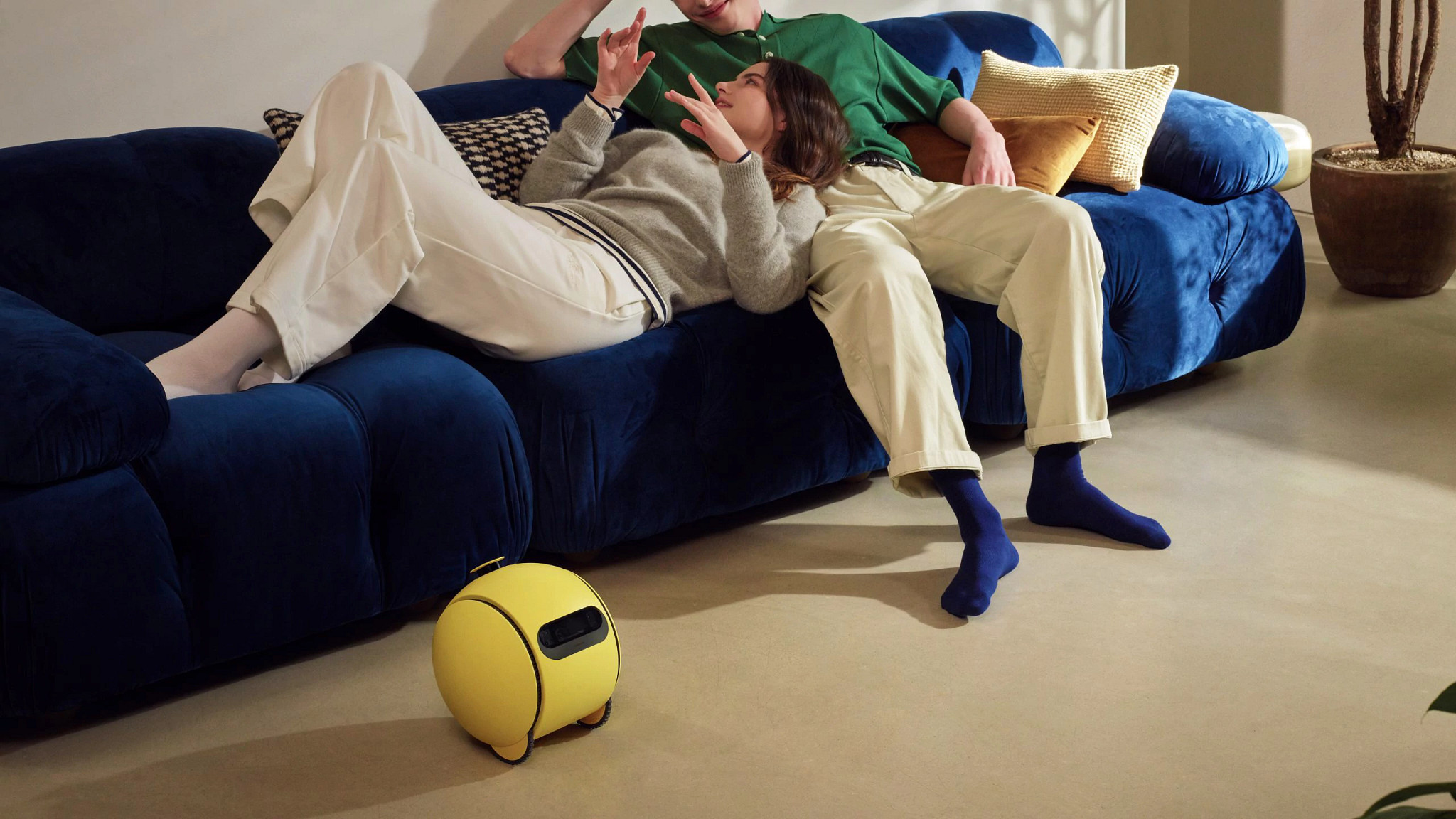 A photo shows a couple louging on a couch. On the floor in front of them is the Samsung Ballie 