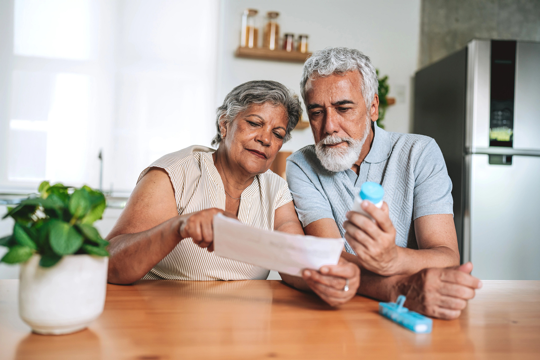 A senior couple sitting at the kitchen table reading a medical prescription or instruction leaflet. The husband is holding a pill bottle while his wife reads the document, as they focus on managing their health and medication routine together at home