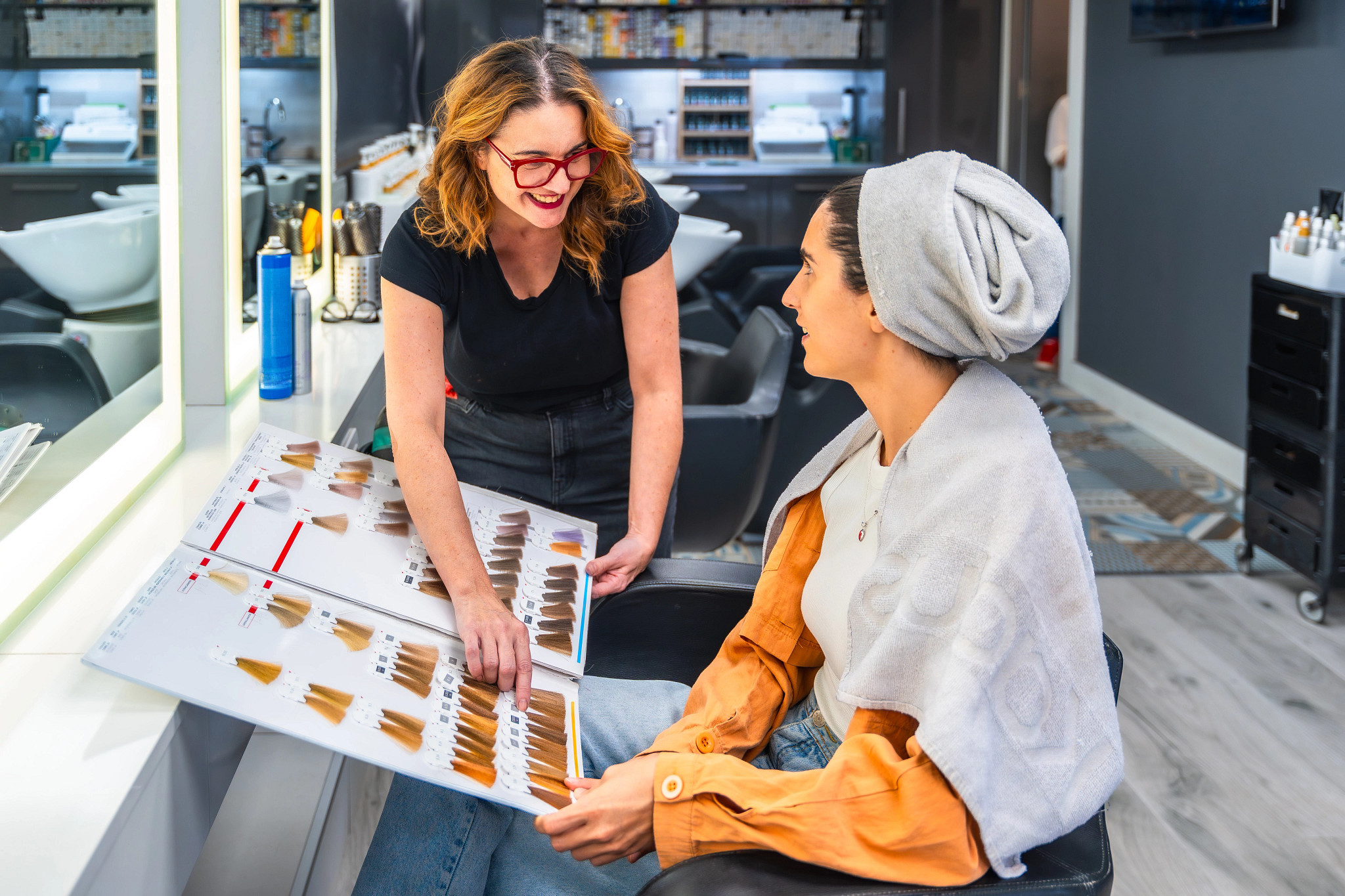 A salon client seated in a styling chair reviews a hair color swatch book with a beauty professional in a modern salon.
