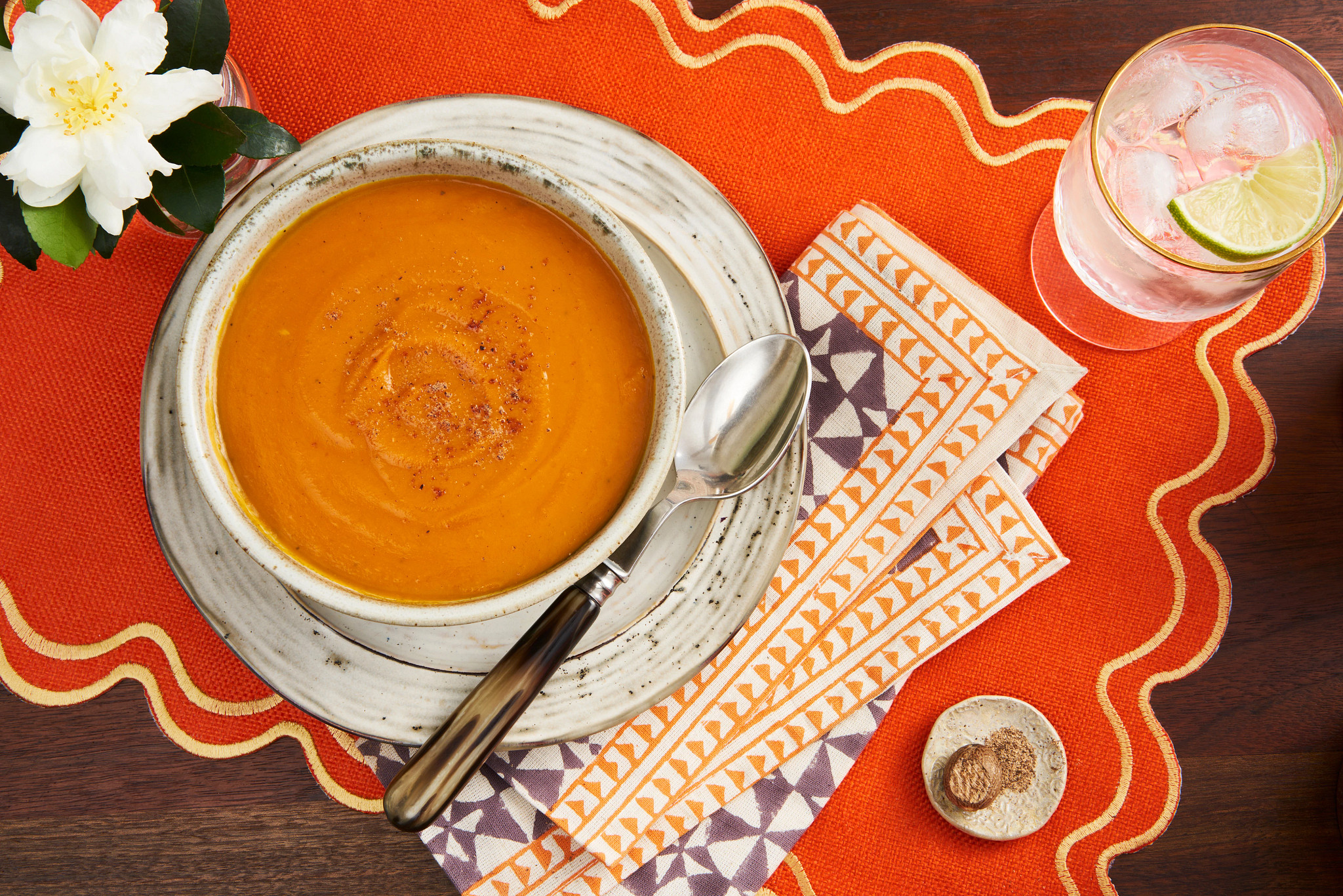 Creamy orange holiday soup served in a white bowl on a festive orange placemat beside a cocktail and white flower