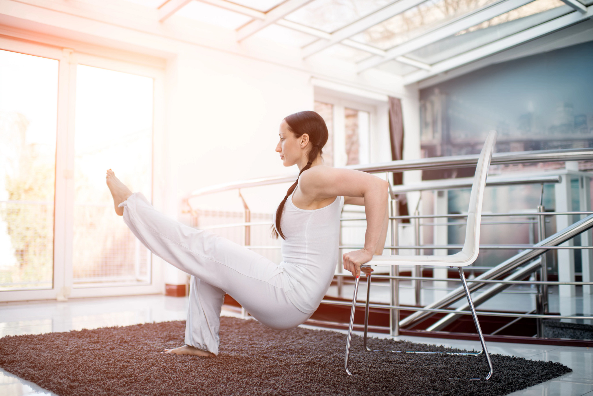 Woman dressed in white performing exercise inside her home with a chair. Window with bright light is in the background.