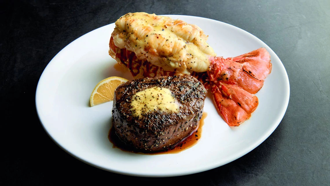 Chart House Butterflied lobster tail next to filet steak topped with butter, lemon wedge on side, all on top of white plate, dark gray background
