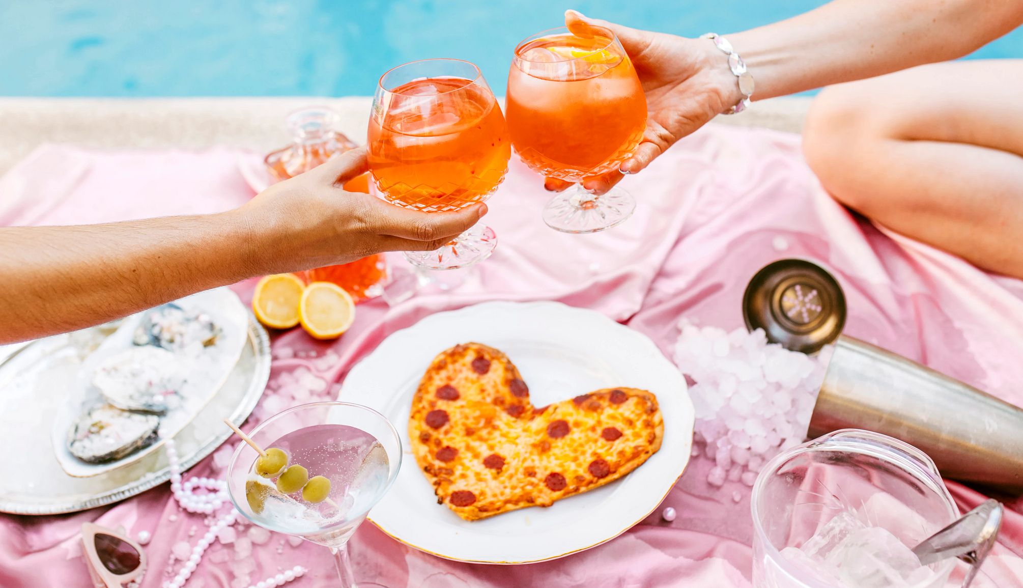 Food and Drink Deals for Mom A photo of a couple toasting cocktails atop a pink blanket with a heart-shaped pizza and a martini.