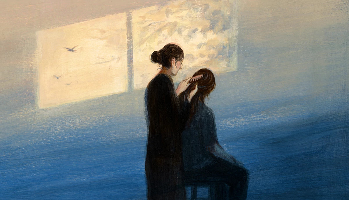 One person gently brushing another person's hair while seated beside a sunlit window.