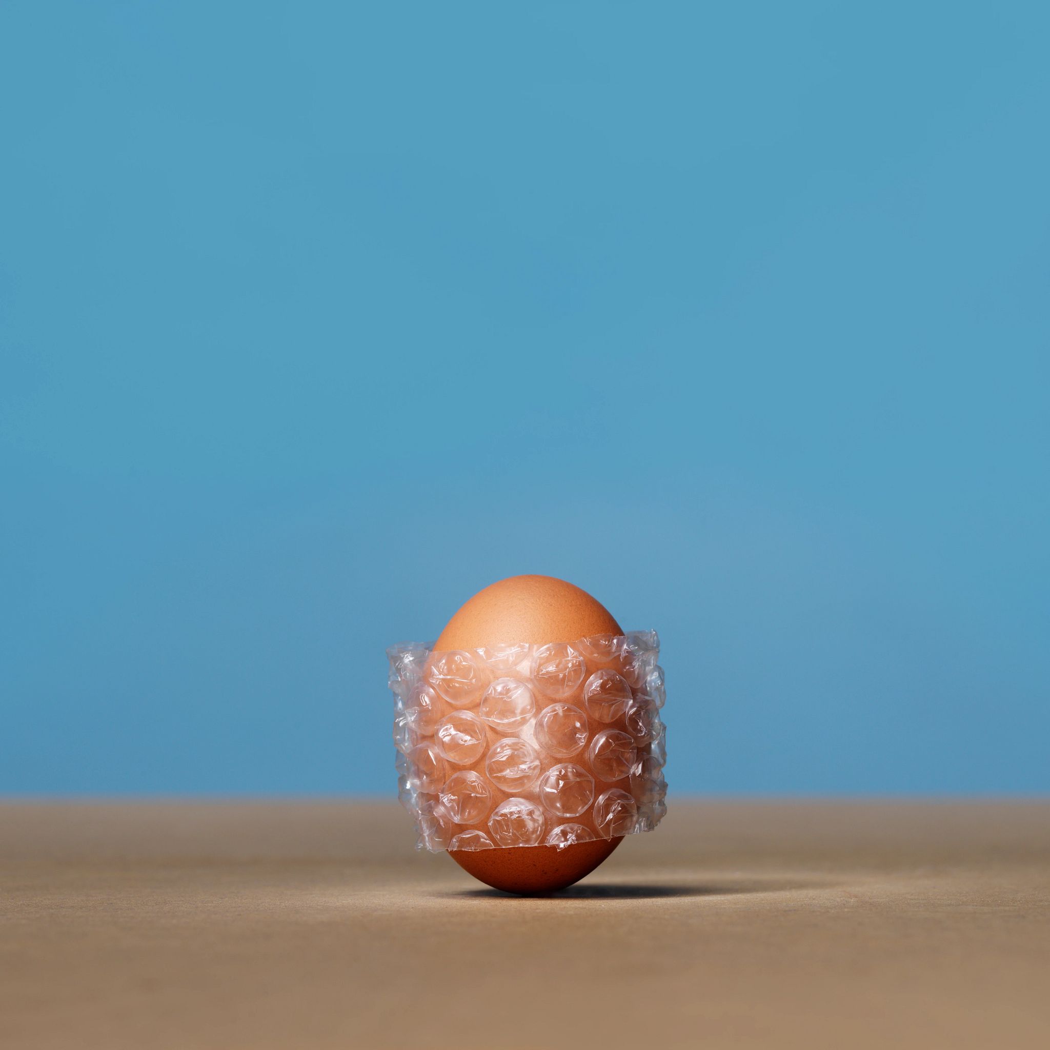 brown egg stands upright on a flat surface against a solid blue background, wrapped securely in a strip of clear bubble wrap to symbolize fragility and protection