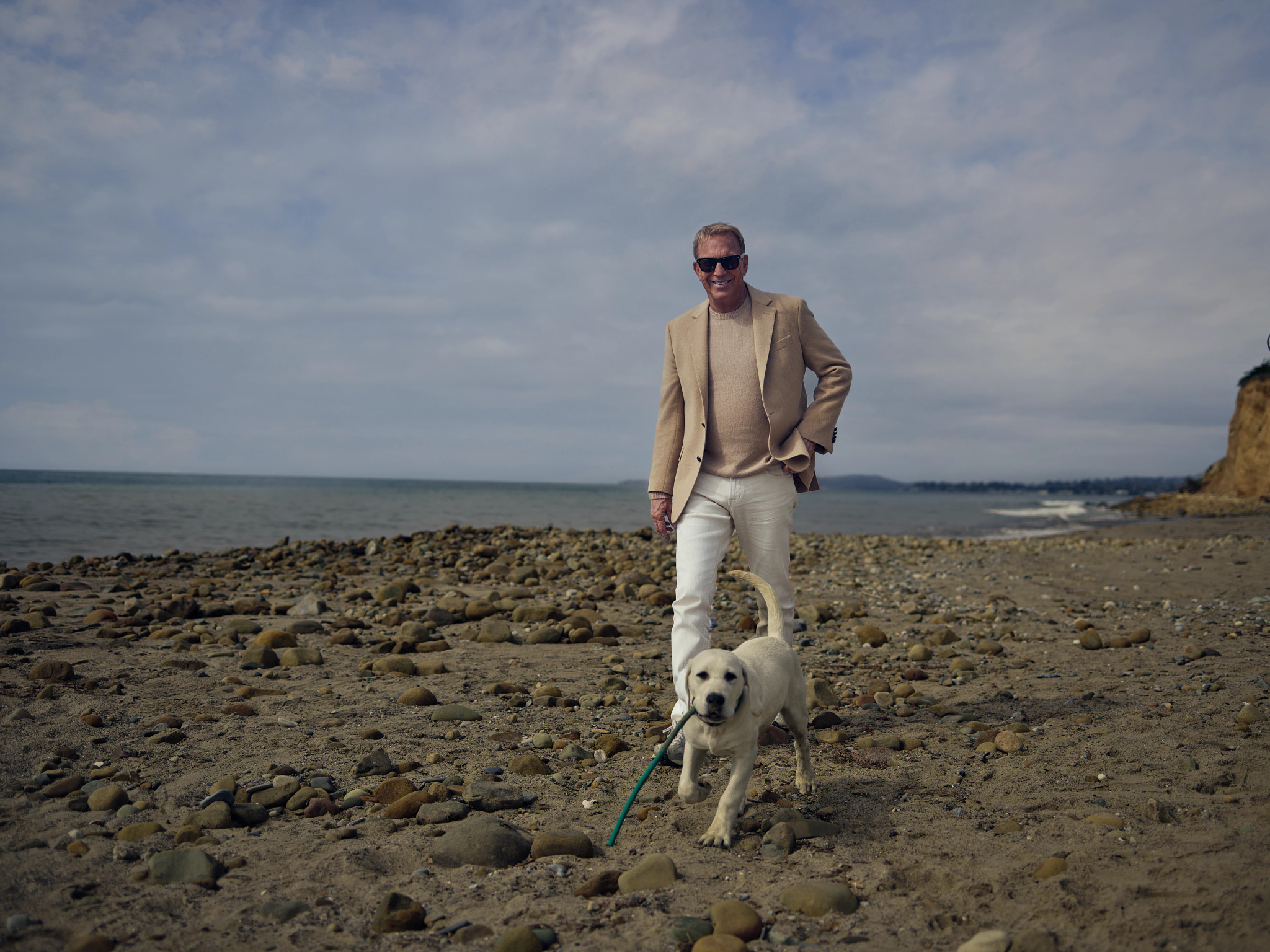 Kevin Costner caminando junto a un perro con el mar de fondo