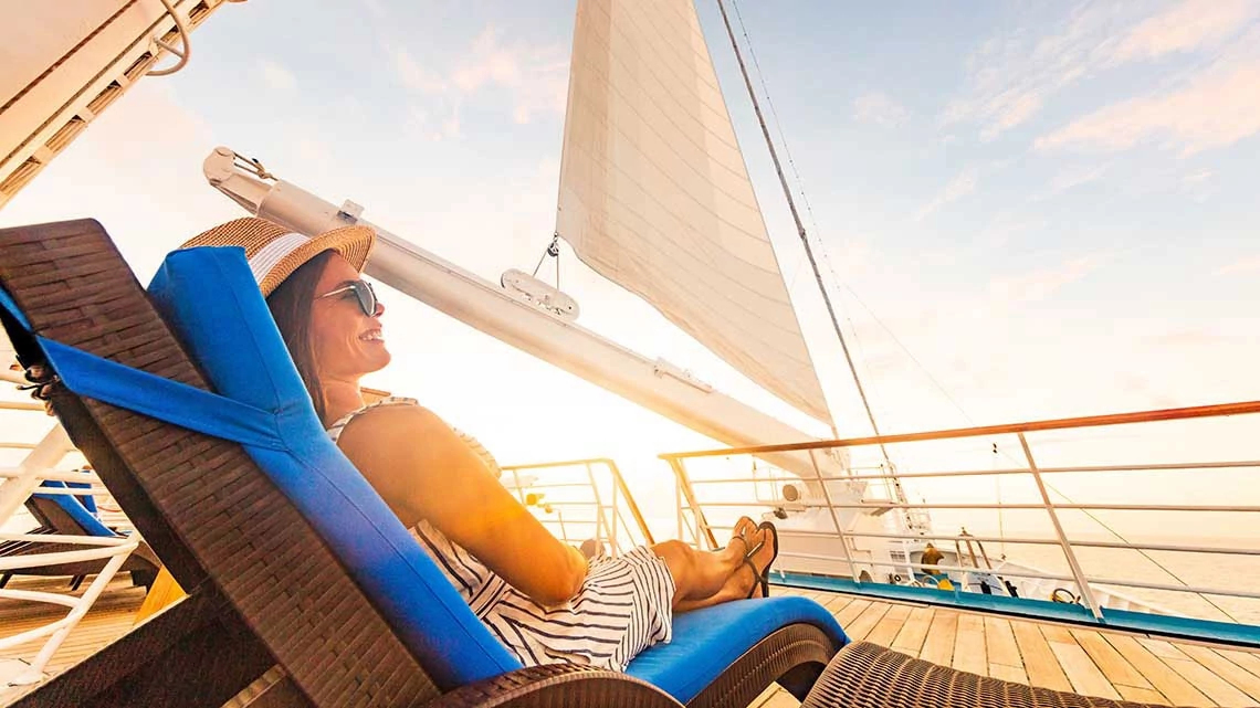 Luxury cruise vacation woman relaxing in lounger chair enjoying sunset on yacht deck with sail in wind sailing in getaway destination summer travel lifestyle.