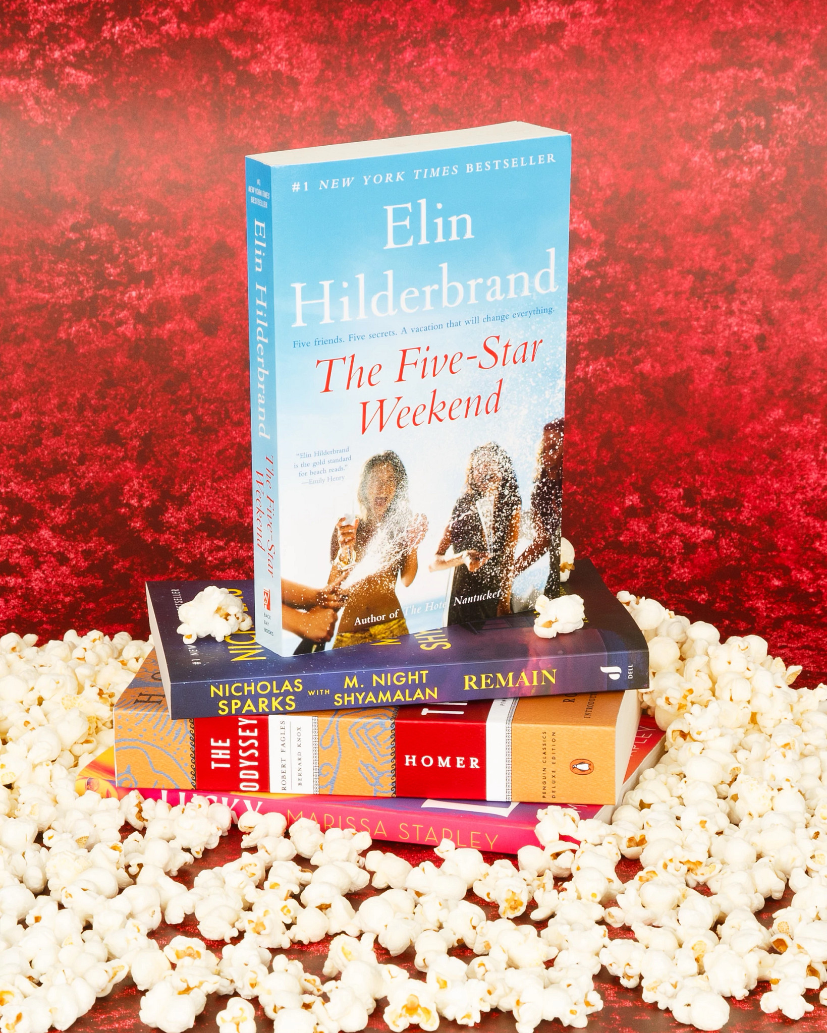A colorful stack of popular books topped by The Five-Star Weekend by Elin Hilderbrand, styled with scattered popcorn against a vibrant red backdrop