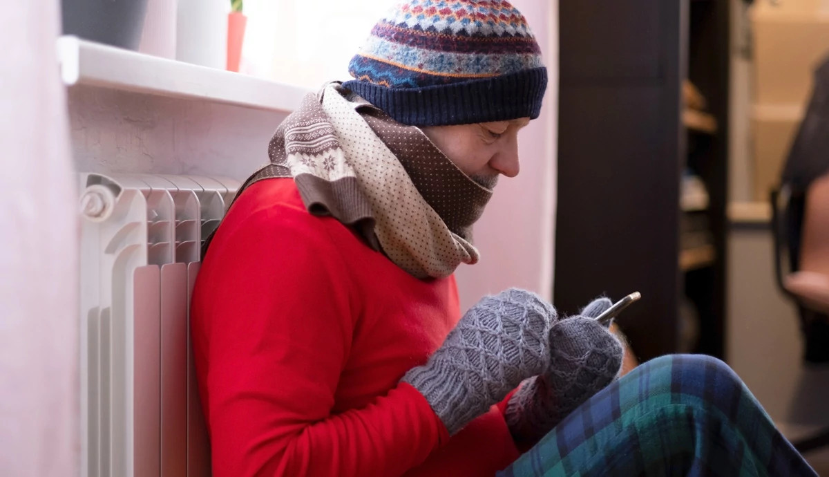 A person wearing a coat, scarf, and gloves and sitting against a home heating unit as they use their cell phone.