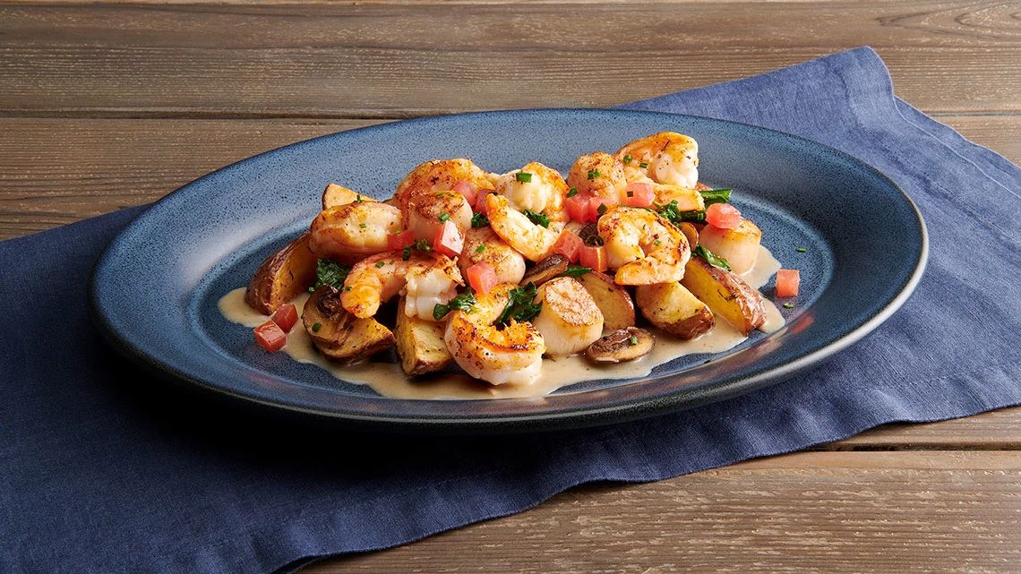 Landry's Seafood wood table, blue napkin, blue plate with scallops, shrimp, potatoes, sauce, tomatoes, parsley