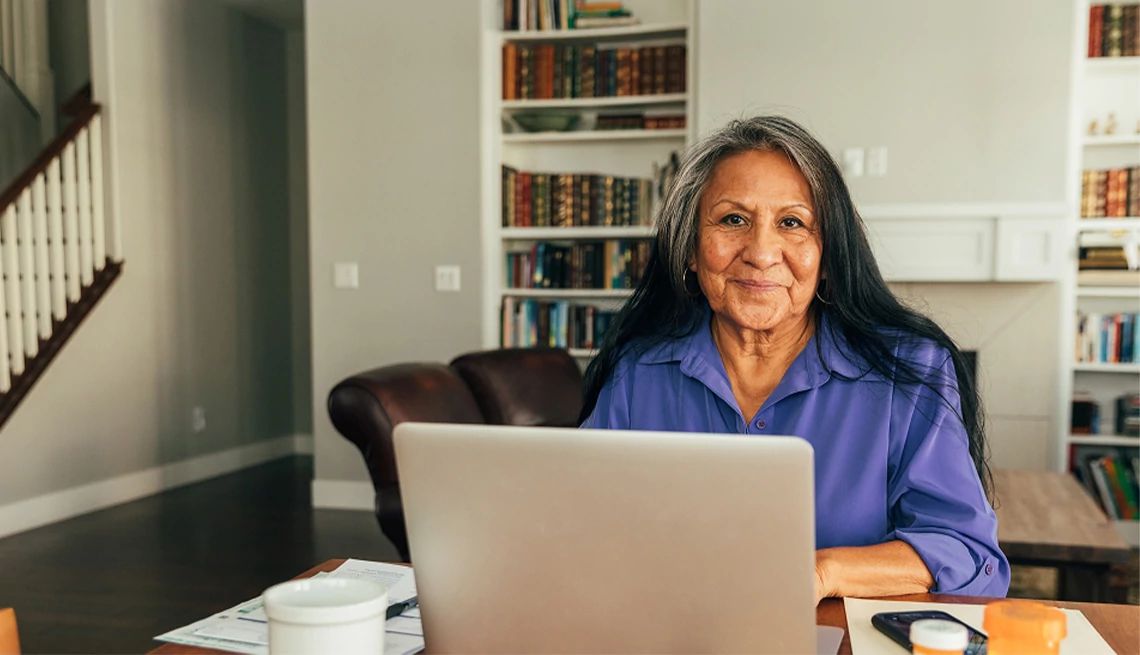Mature woman sitting at desk in her home office looking at AARP Medicare resources on her laptop