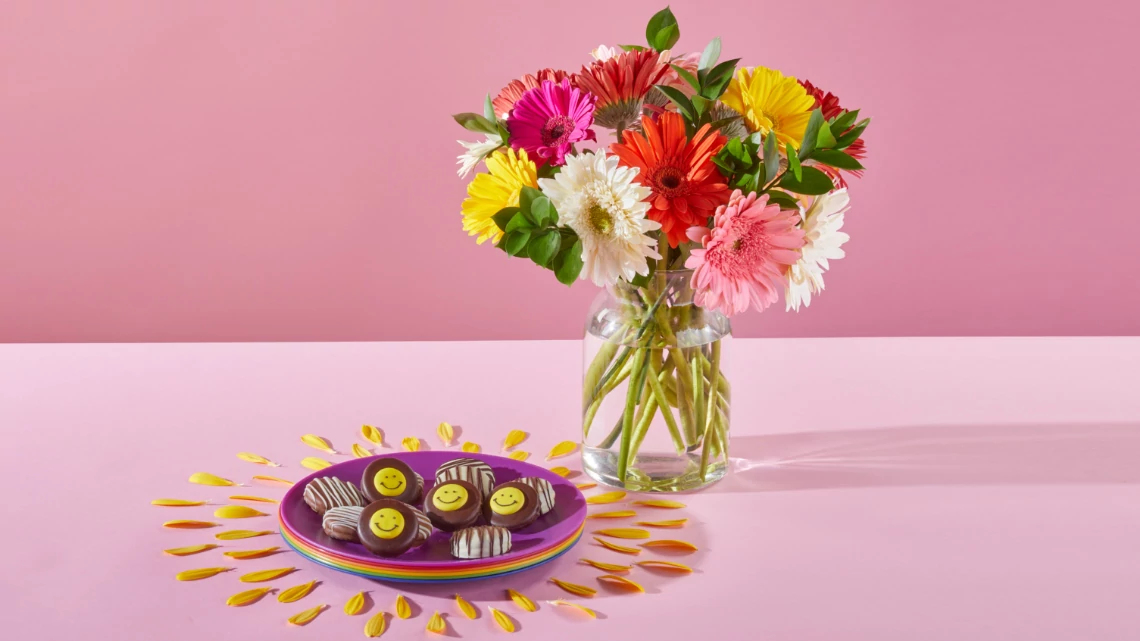 red, pink, yellow, white gerber daisy flowers in a clear glass vase on a pink table with purple plate next to it holding yellow smile face chocolate white stripe cookies