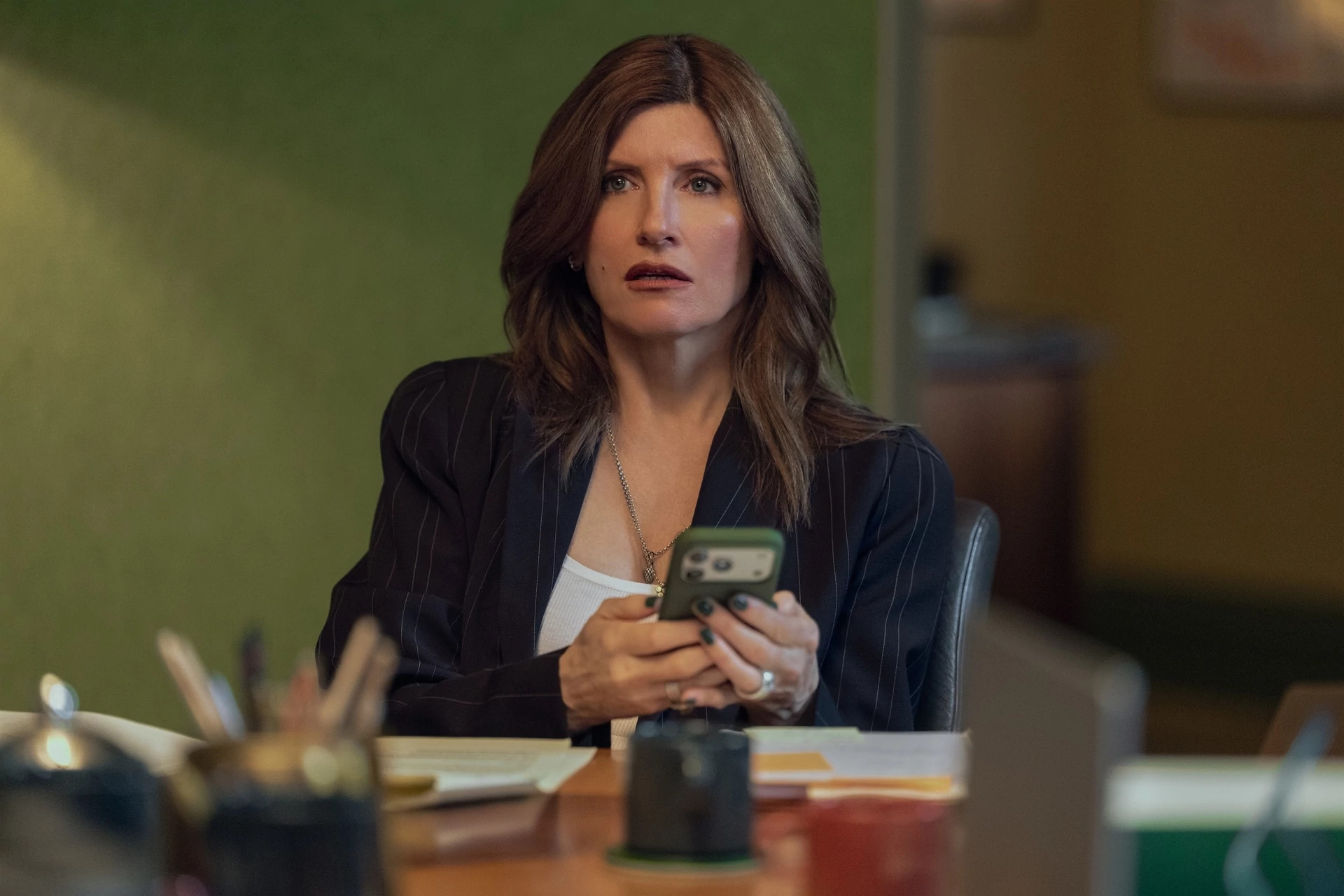 Sharon Horgan seated at an office desk holding a smartphone in the television show, "Youth."