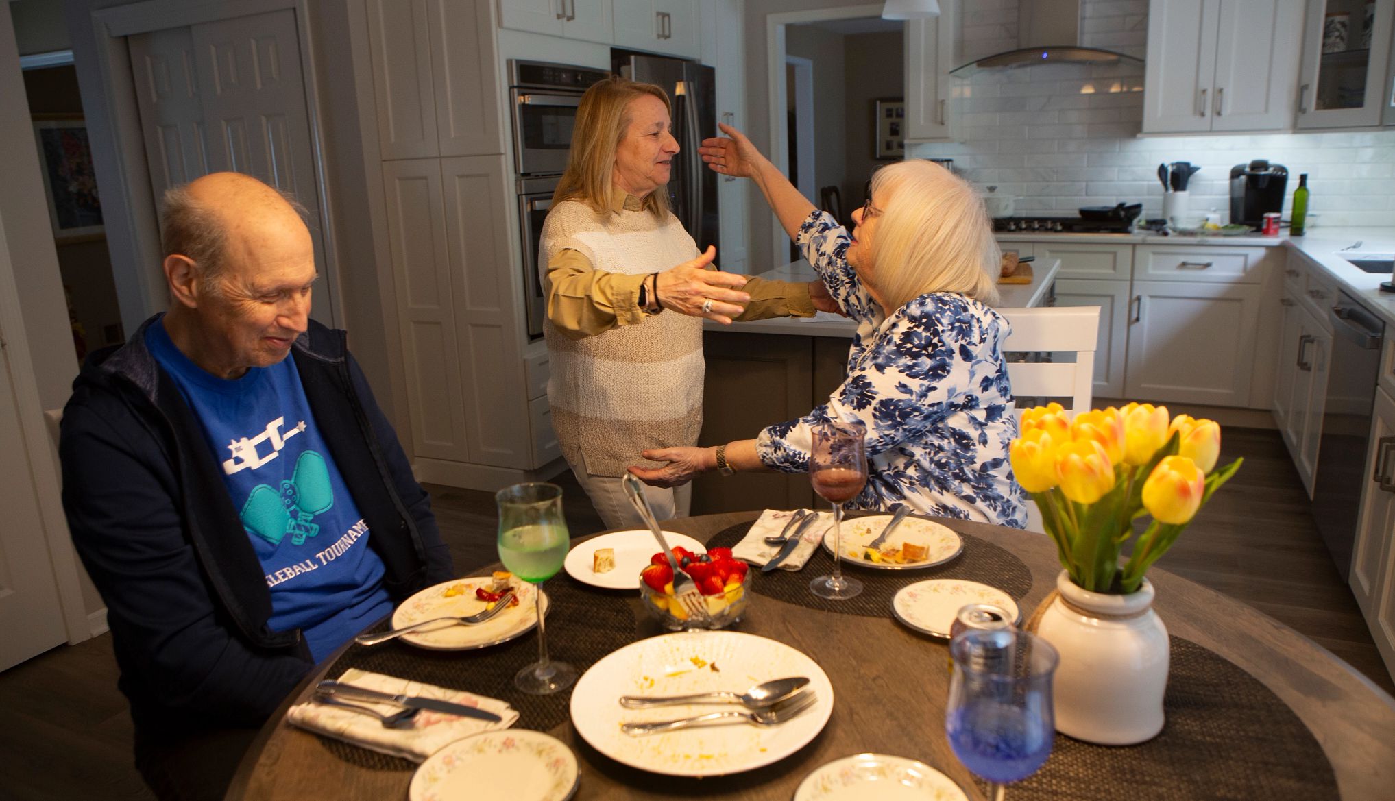 judy rosenstein reaches out to hug her friend, wendy stein. judy and her husband, abe, are seated at a kitchen table with plates of food in front of them.