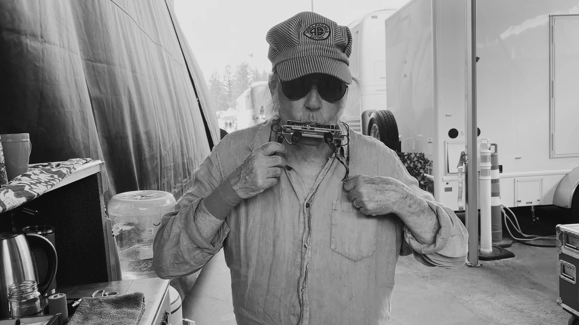 neil young, wearing a conductor's hat and sunglasses, blowing into a harmonica that is on a neck holder