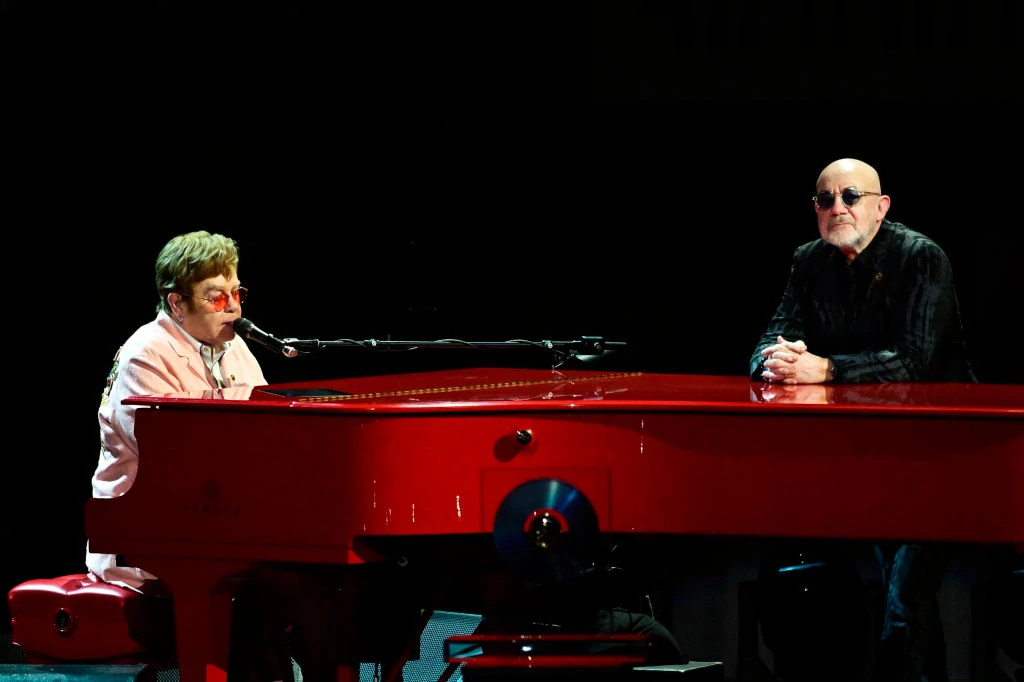 Elton John performing on the piano onstage with Bernie Taupin standing nearby