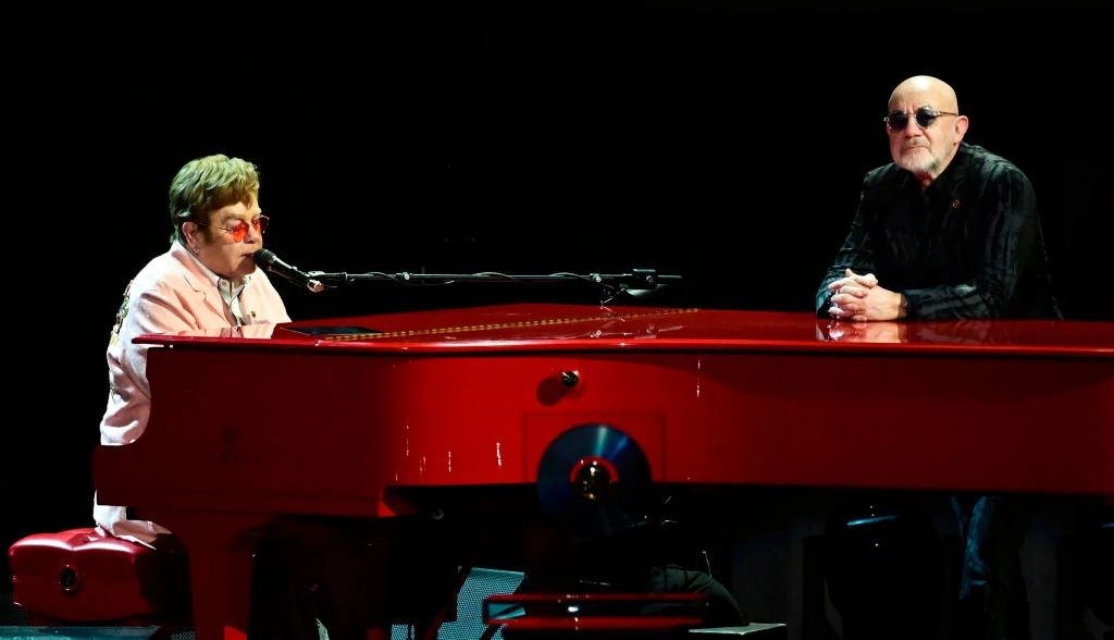 Elton John performing on the piano onstage with Bernie Taupin standing nearby