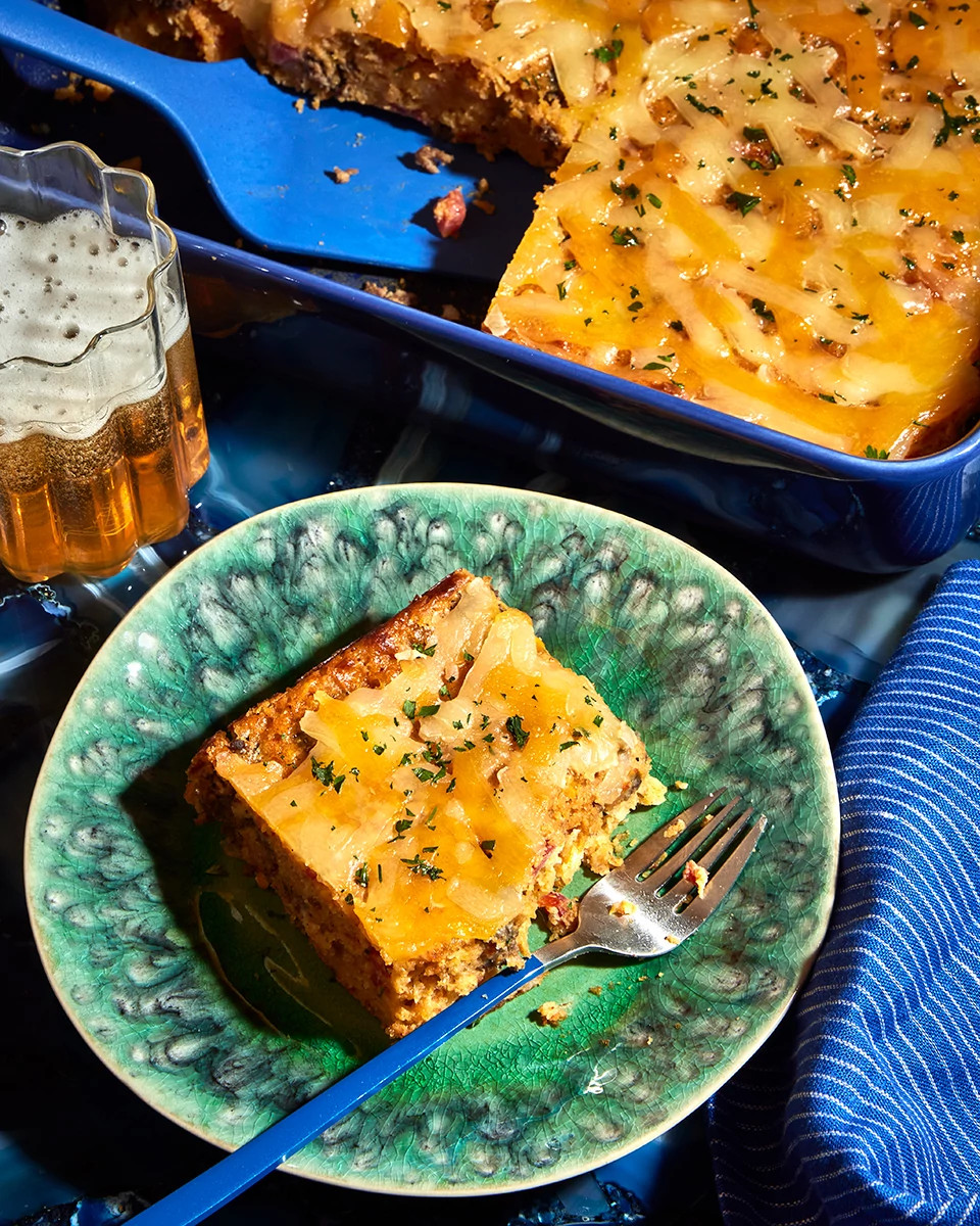 Slice of cheesy casserole on a green plate with fork beside a baking dish and glass of beer