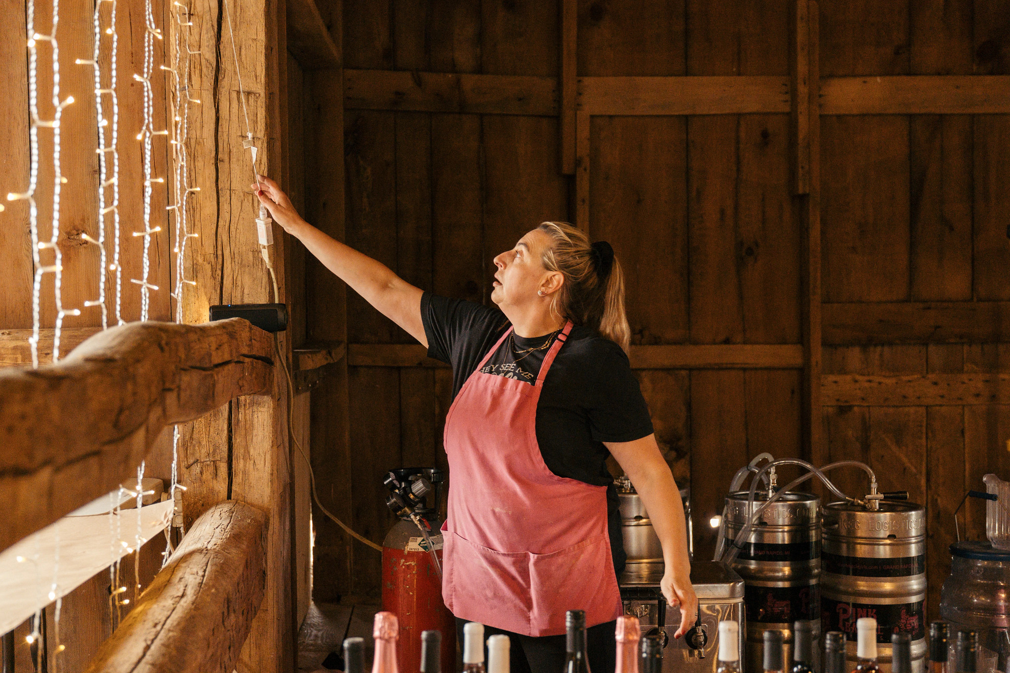 Stephanie Ginsberg inside a rustic wooden workspace adjusting decorative string lights near shelves and beverage equipment
