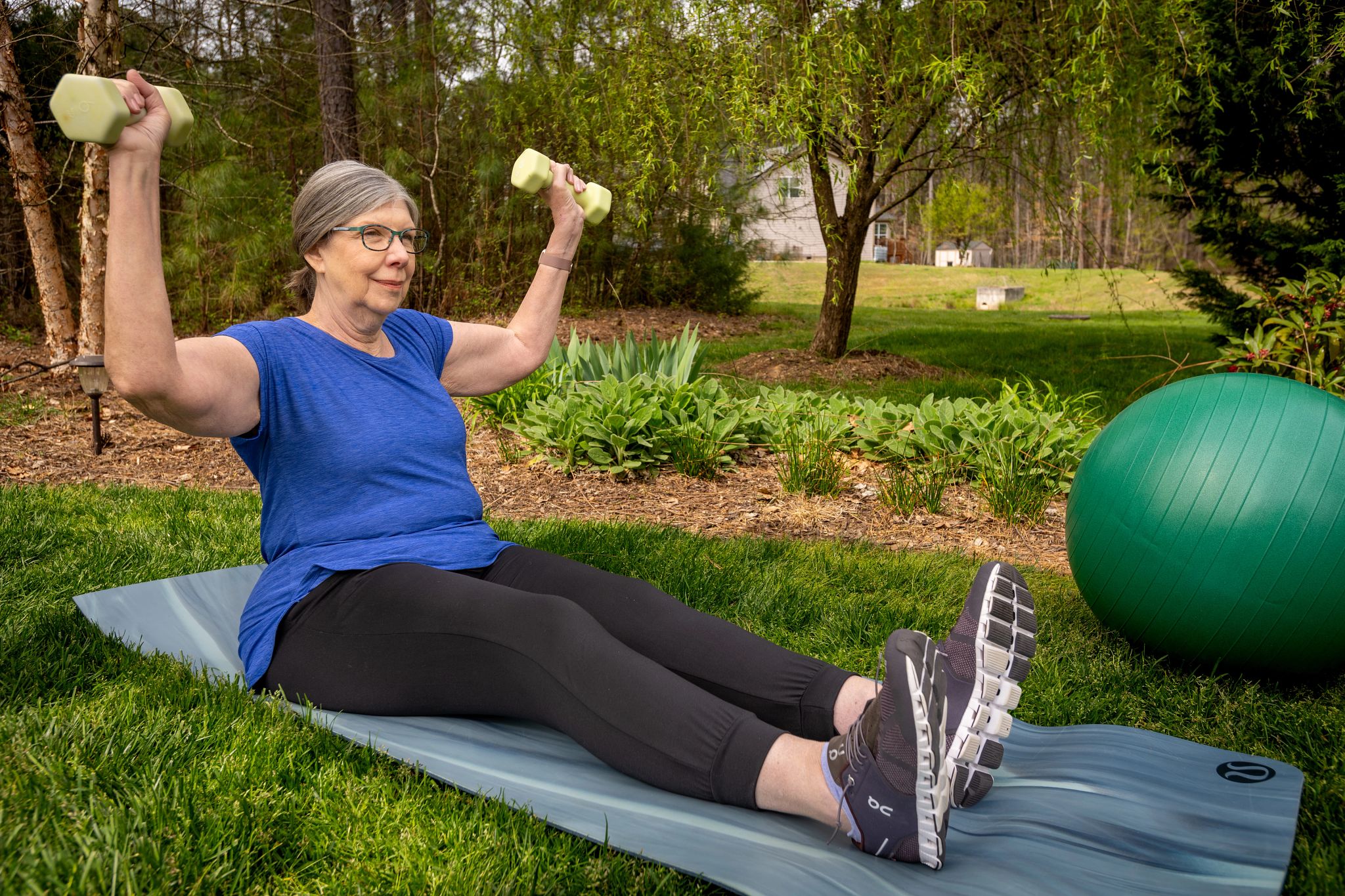 Angela Cheney strength training at her home in Durham, North Carolina Angela Cheney sitting on mat with legs straight out, lifting dumbbell in each hand, arms out to side at 90 degree angle