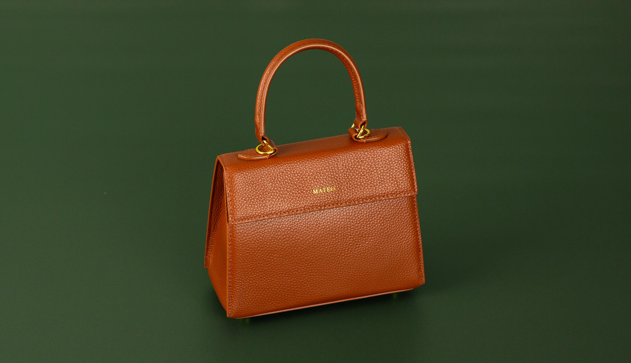Studio product photo of a Mateo leather handbag in warm cognac brown with a structured top-handle silhouette and textured leather finish displayed against a dark green background