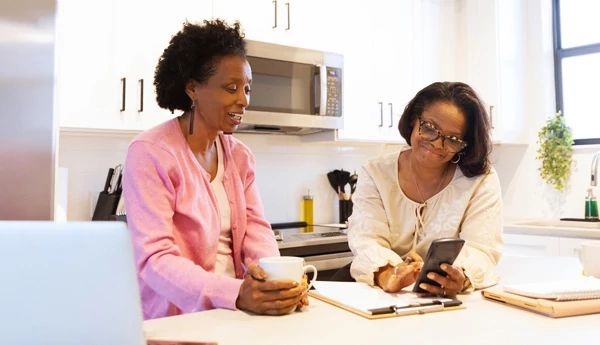 Two women seated in the kitchen looking at a phone one of the women is holding.