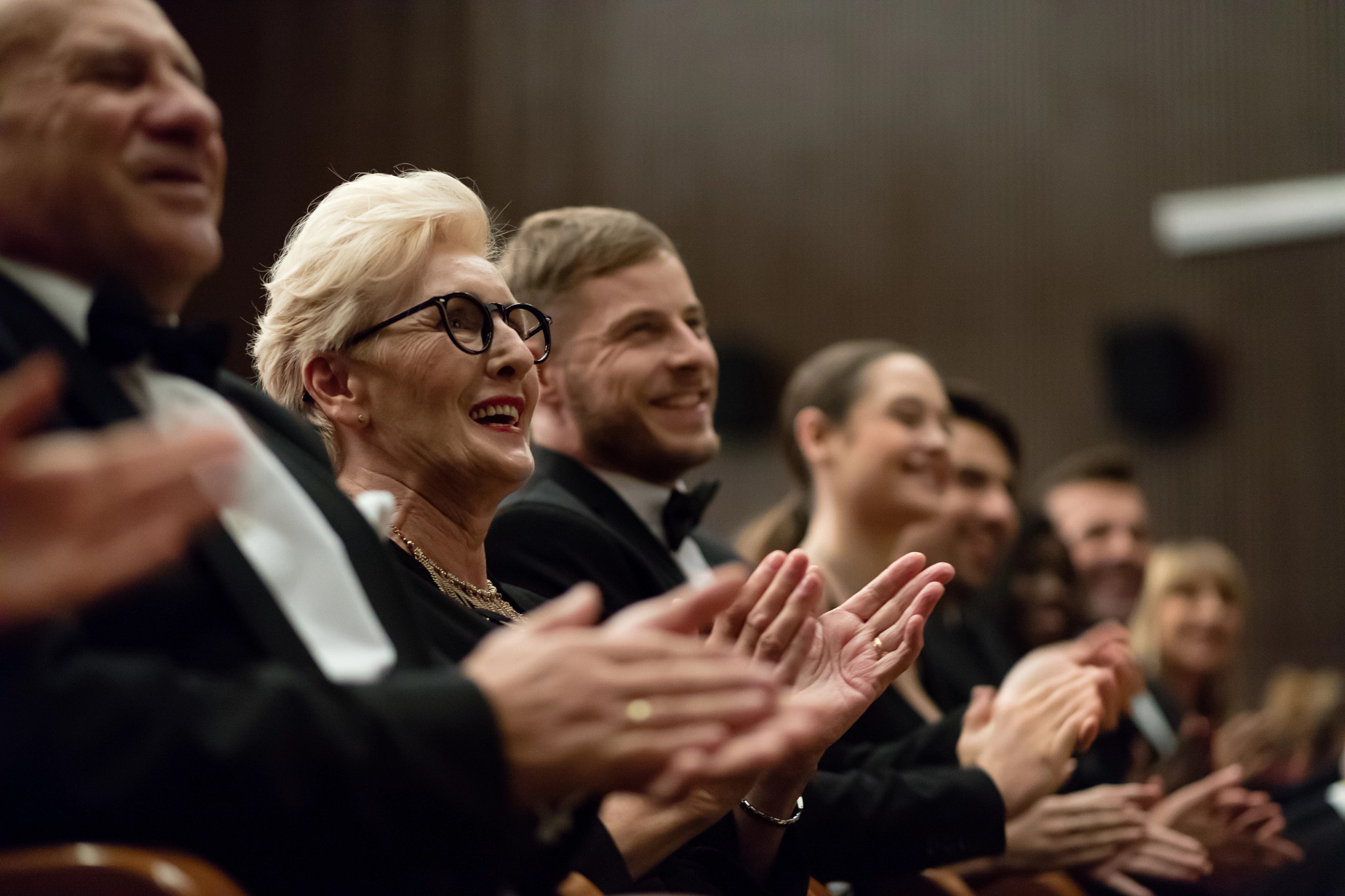 Side view of audience clapping hands in opera house. Men and women are watching theatrical performance. They are in elegant wear.