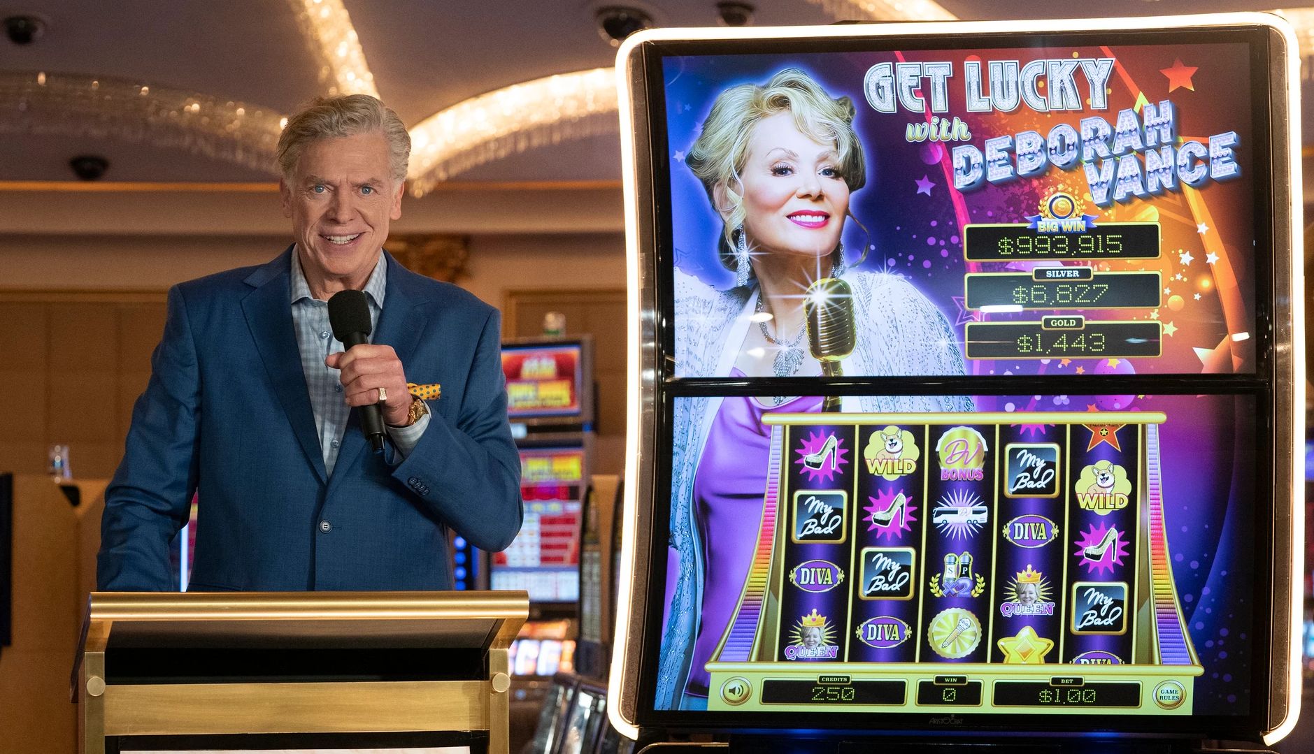 Christopher McDonald holding a microphone standing next to slot machine featuring Jean Smart's character Deborah Vance in Hacks