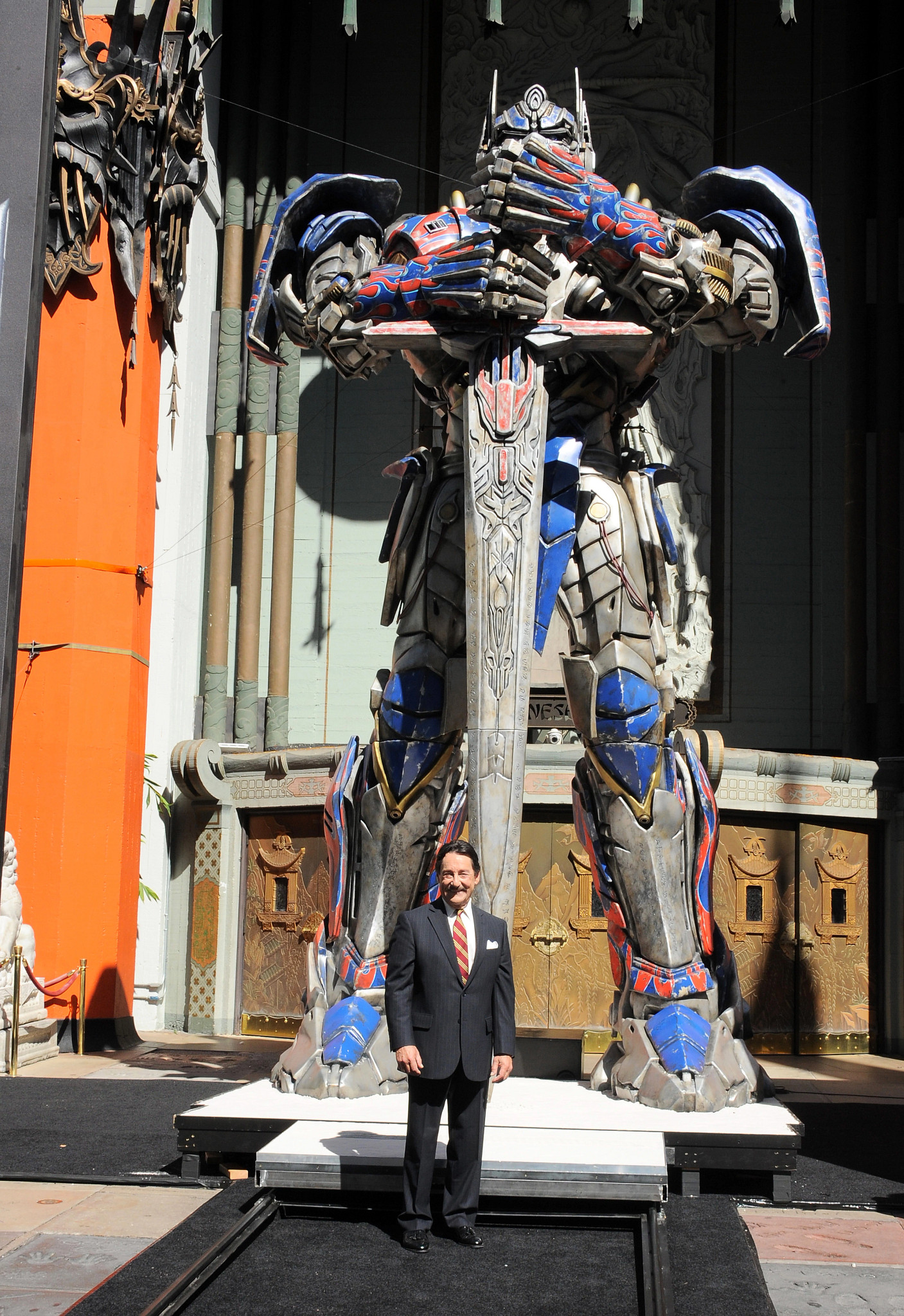 peter cullen in a dress suit at a hollywood premiere standing under a huge statue of optimus prime