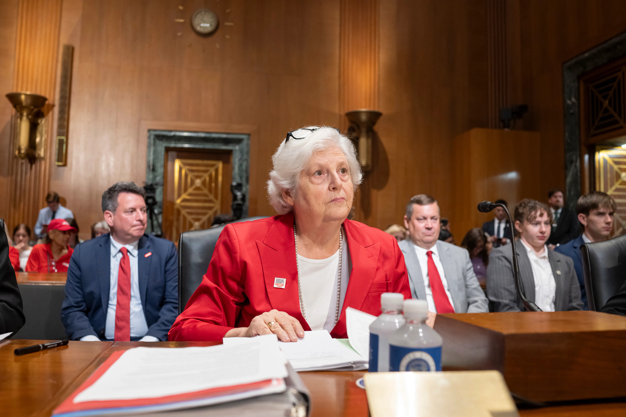 A woman in glasses, a red blazer and white shirt, sits in front of a microphone.