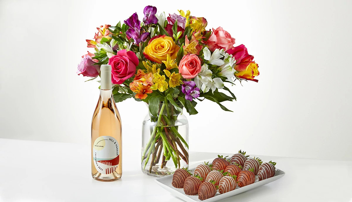 A bouquet of flowers, a bottle of wine, and a plate of chocolate strawberries from FTD sitting on a white table