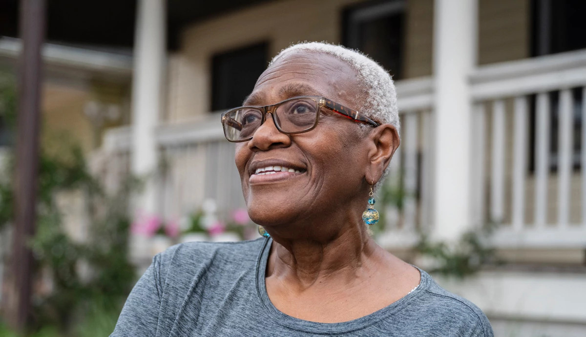 A close-up photo of a short haired woman wearing eyeglasses and a casual top outdoors in front of her home, looking away with a contented smile.