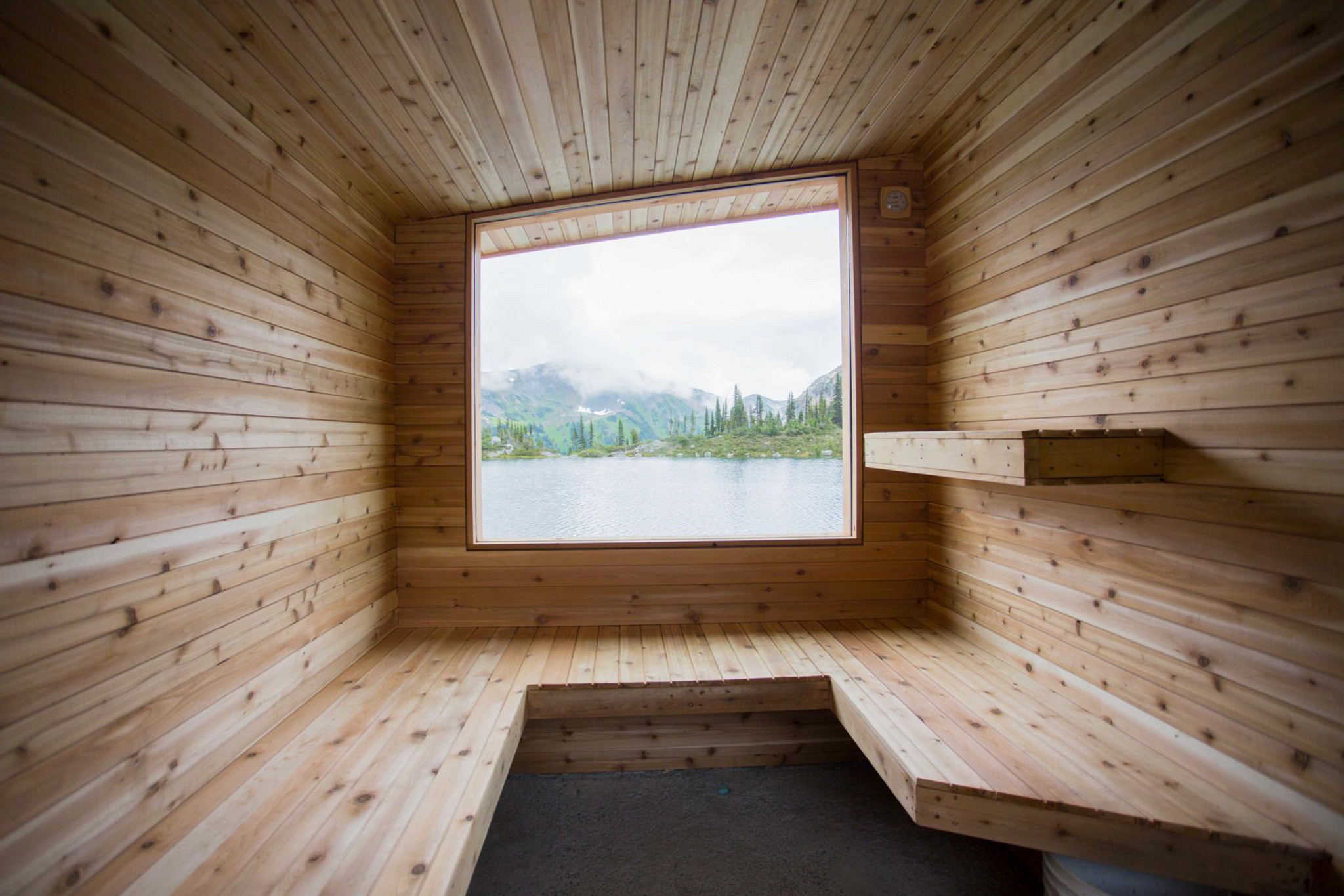 The wood-paneled interior of an empty sauna features a large picture window showing an outdoor view of a lake and mountains under a cloudy sky.
