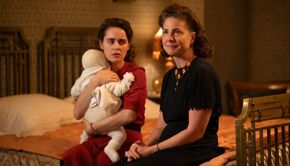 Hadas Yaron holding on a baby while sitting next to Robin Weigert on a bed in We Were the Lucky Ones