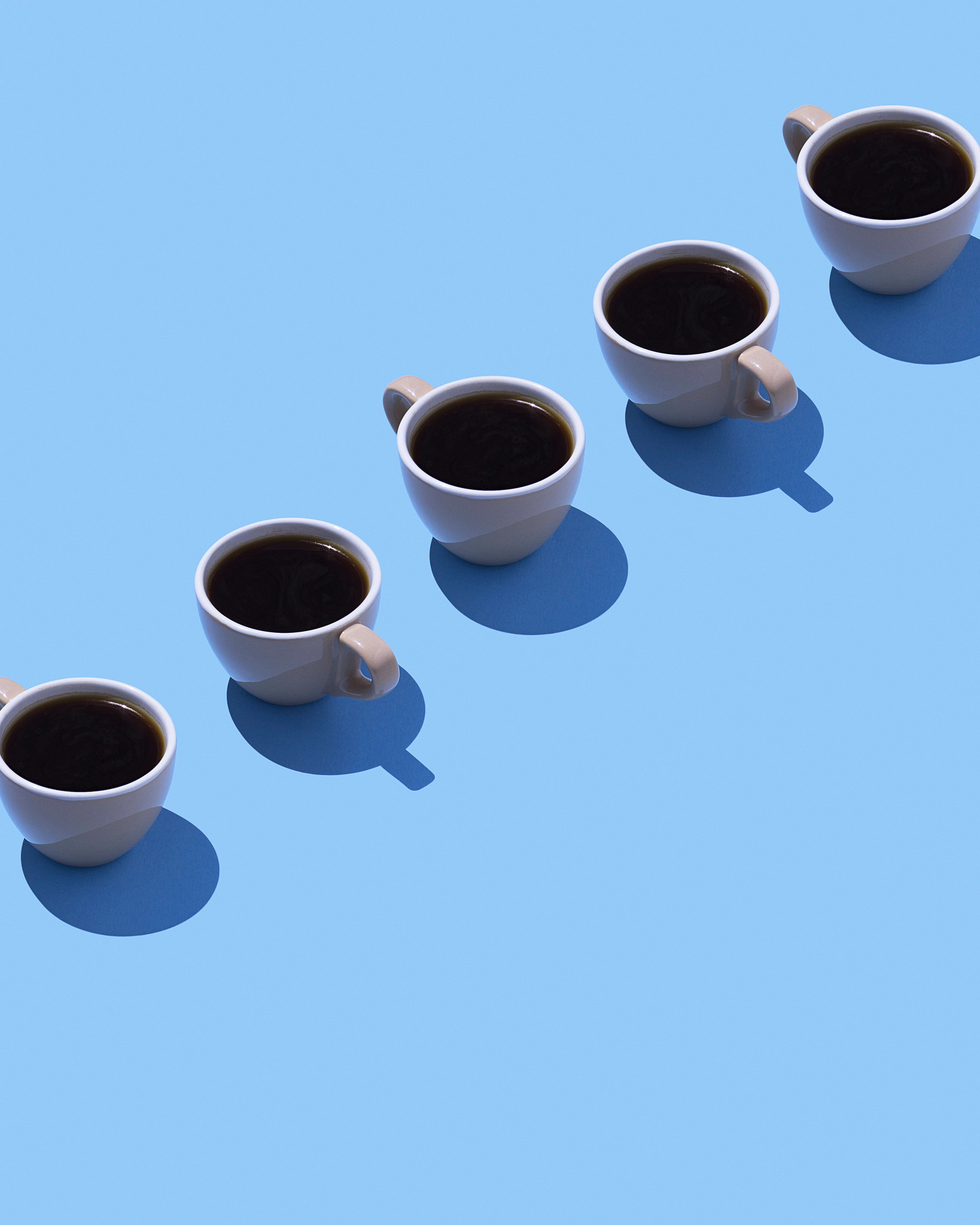 A diagonal row of small ceramic cups filled with black coffee against a bright blue background.