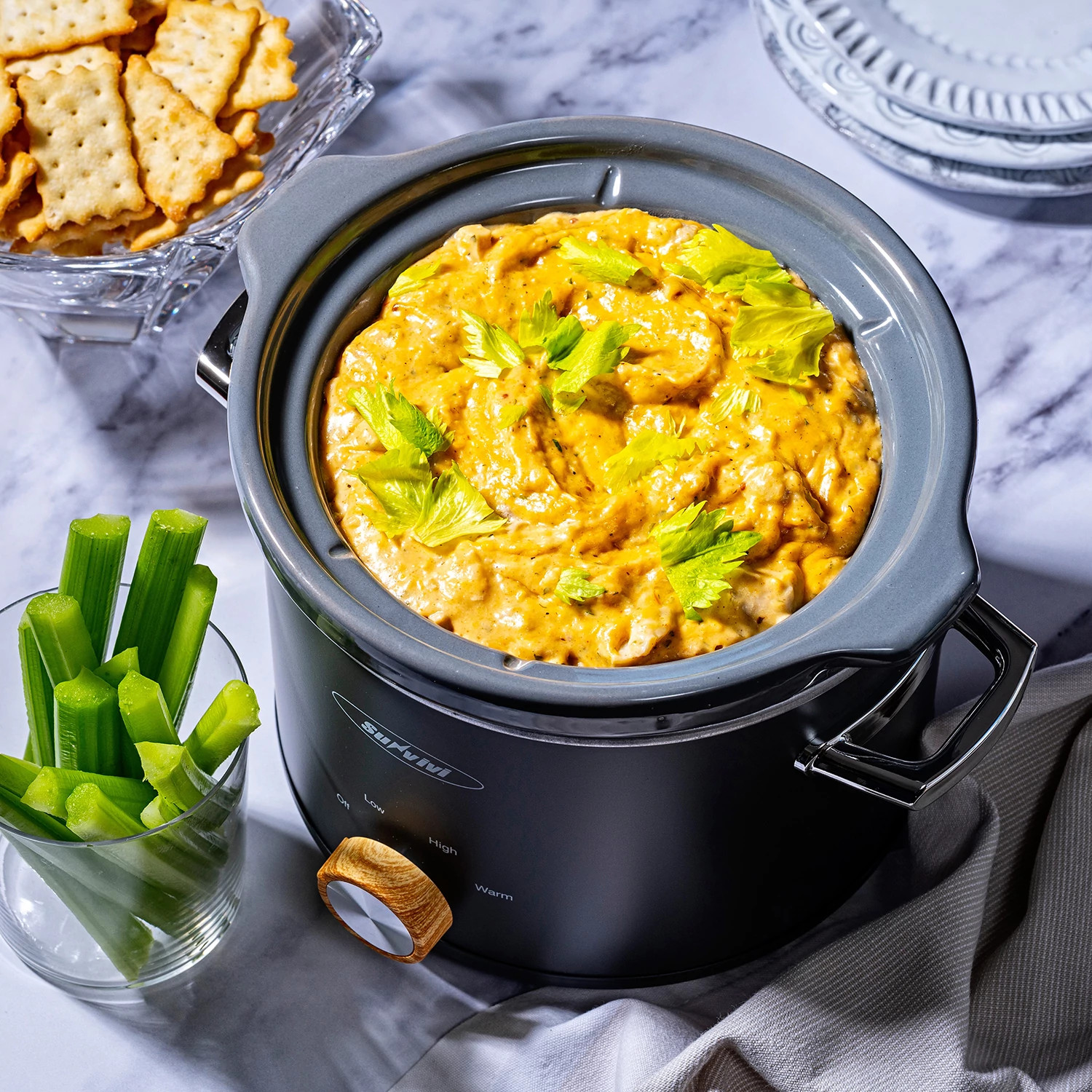 Creamy cheese dip with celery garnish served in a slow cooker alongside crackers and celery sticks.