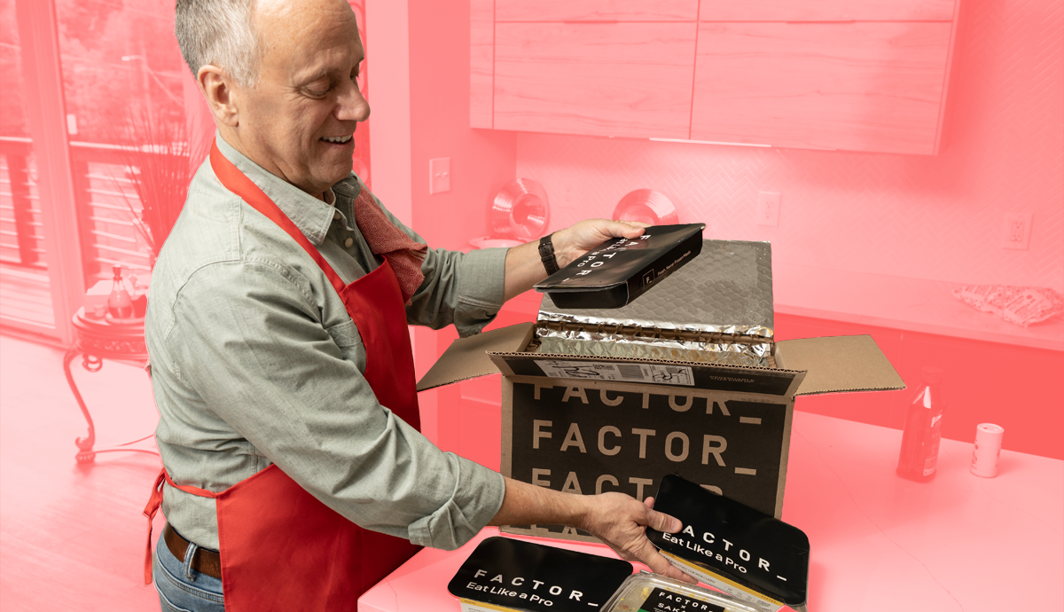 A man wearing an apron opening a box of Factor meals on the kitchen counter with a red background