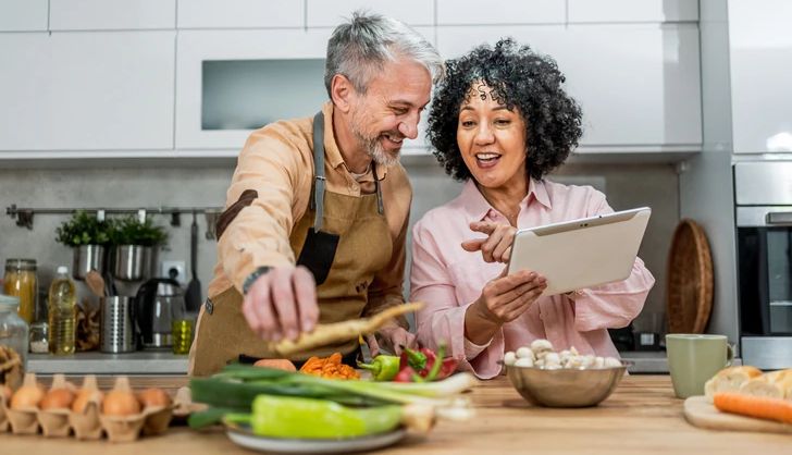 Cheerful mature couple using a digital tablet to look up a recipe while cooking together in the kitchen Cheerful mature couple using a digital tablet to look up a recipe while cooking together in the kitchen
