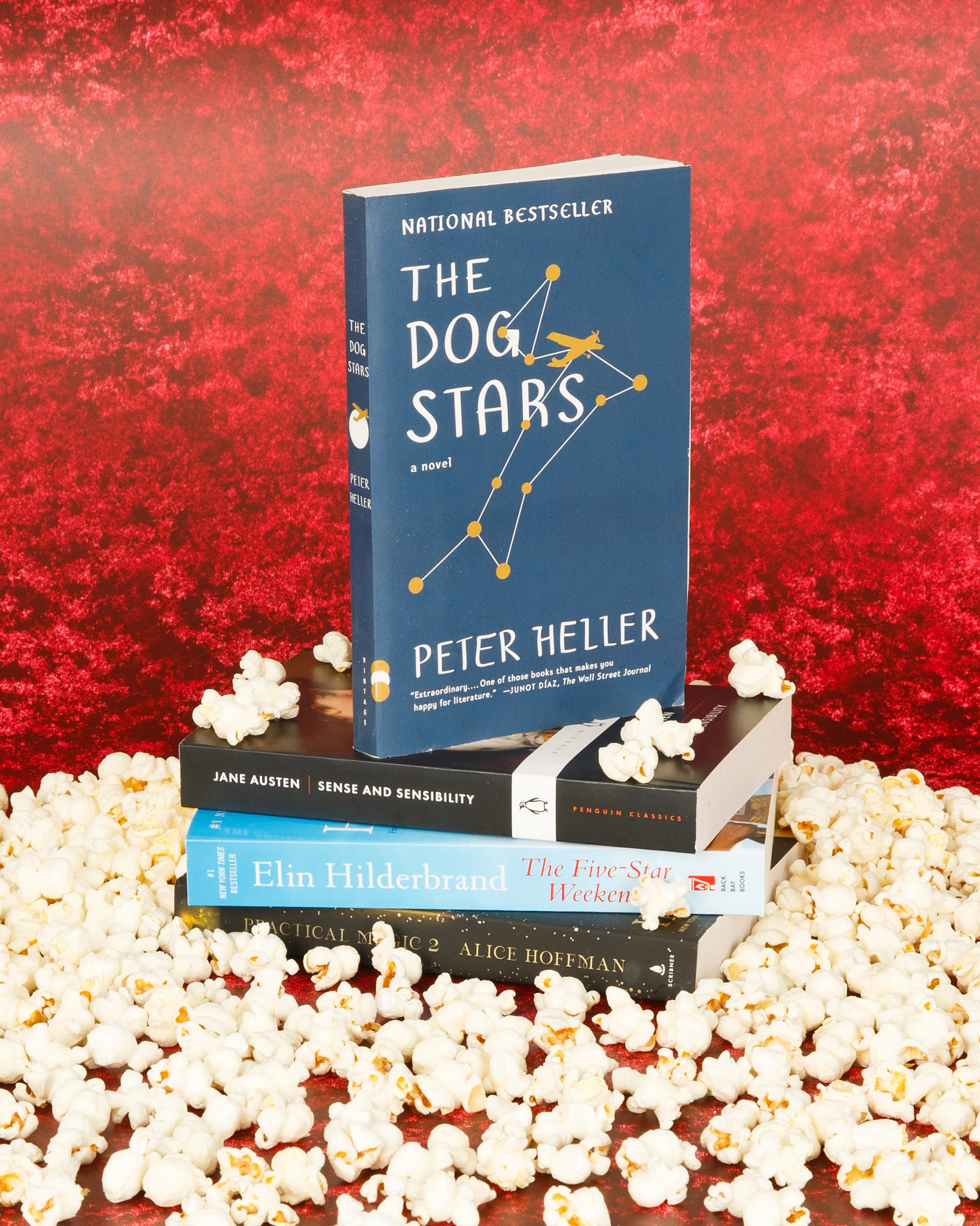 A styled still life featuring the novel “The Dog Stars” by Peter Heller displayed upright on a stack of books including classic and contemporary titles, surrounded by scattered popcorn on a red textured background