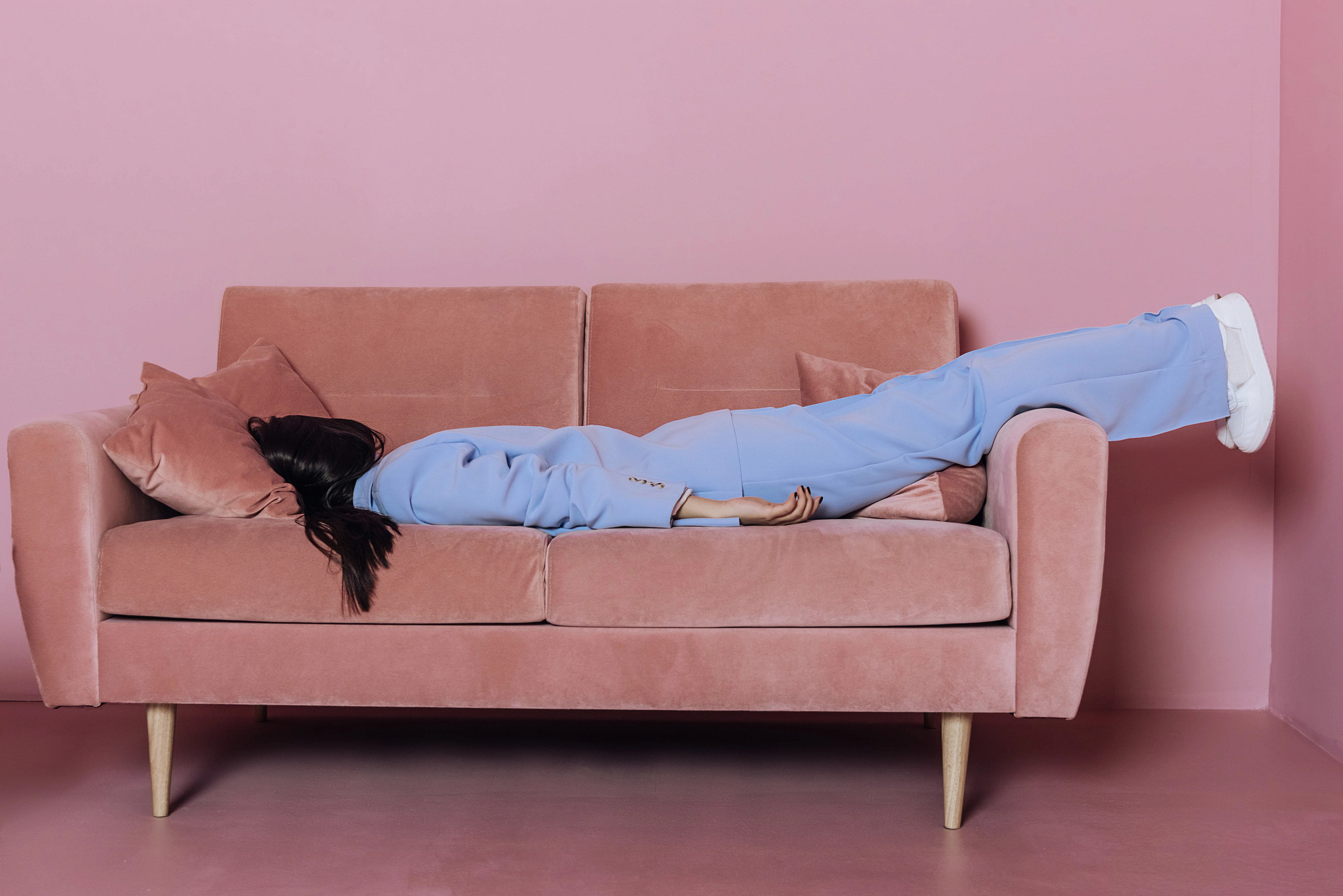 woman in a light blue suit lies face down and exhausted across a dusty-rose-colored sofa against a matching pink wall