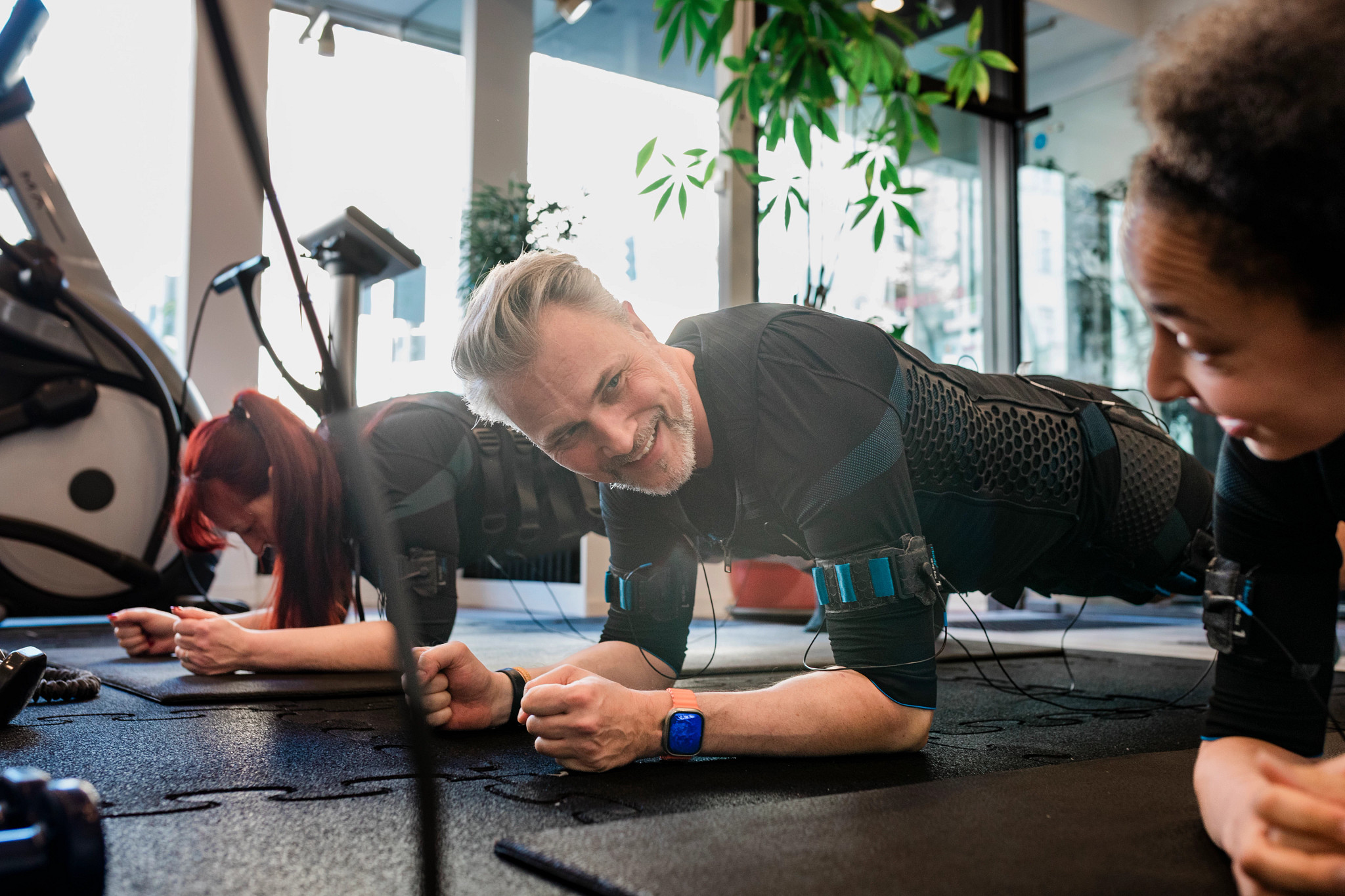 An older adult man and two women wearing electronic muscle stimulation suits while holding a plank position on mats at a gym.