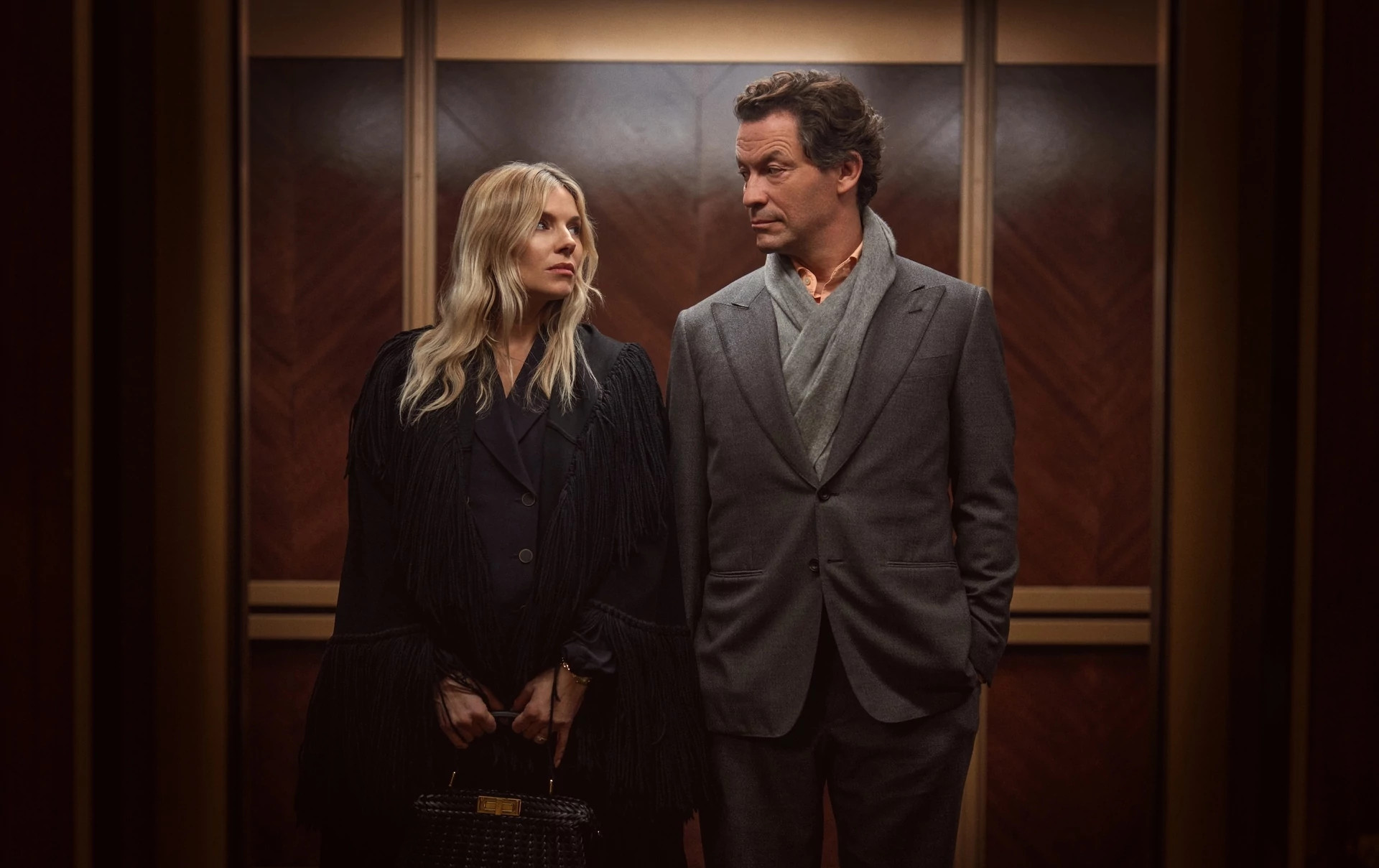 Sienna Miller and Dominic West standing inside a wood-paneled elevator in the television show, "War."