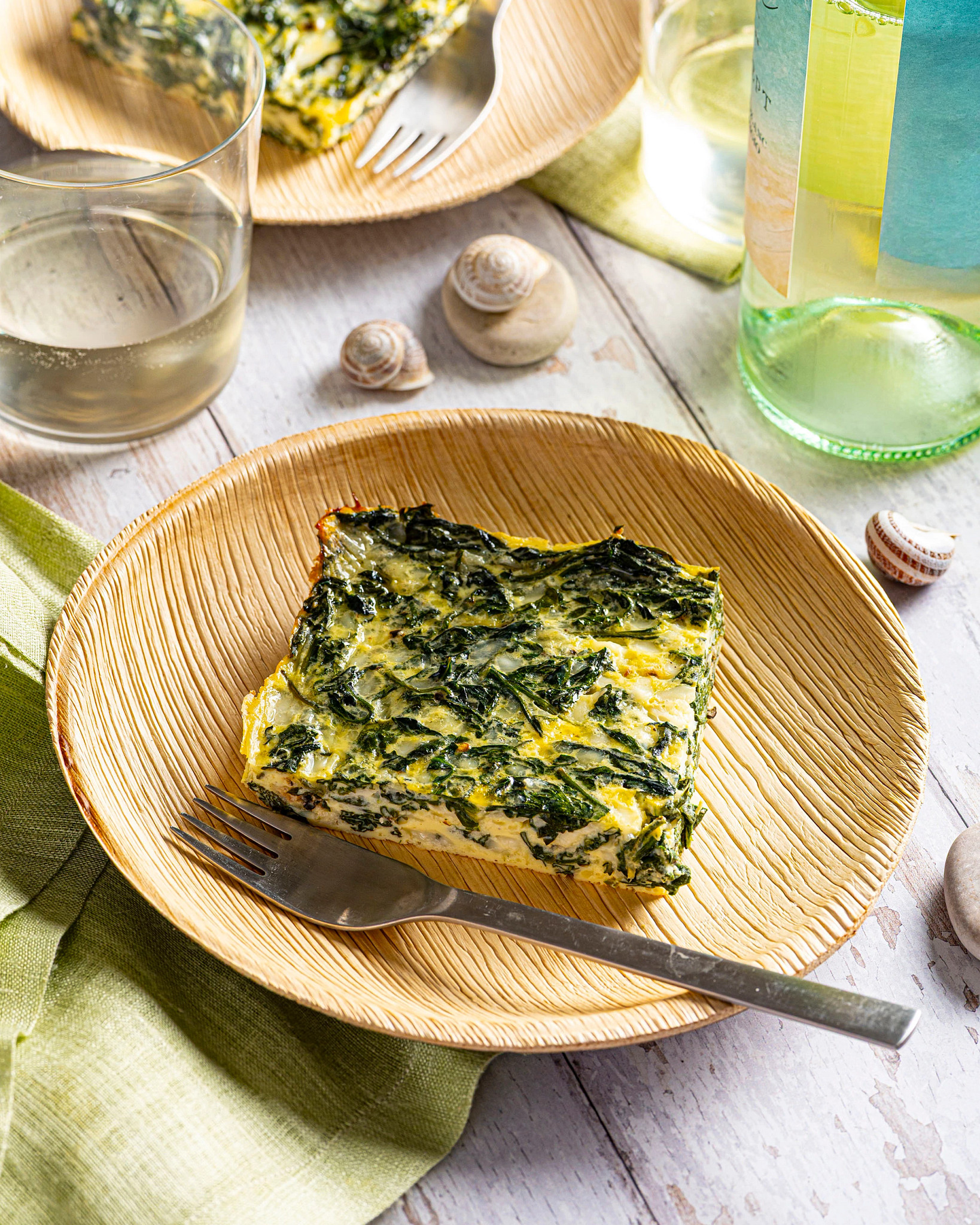 A slice of crustless spinach quiche served on a wooden plate alongside a beverage