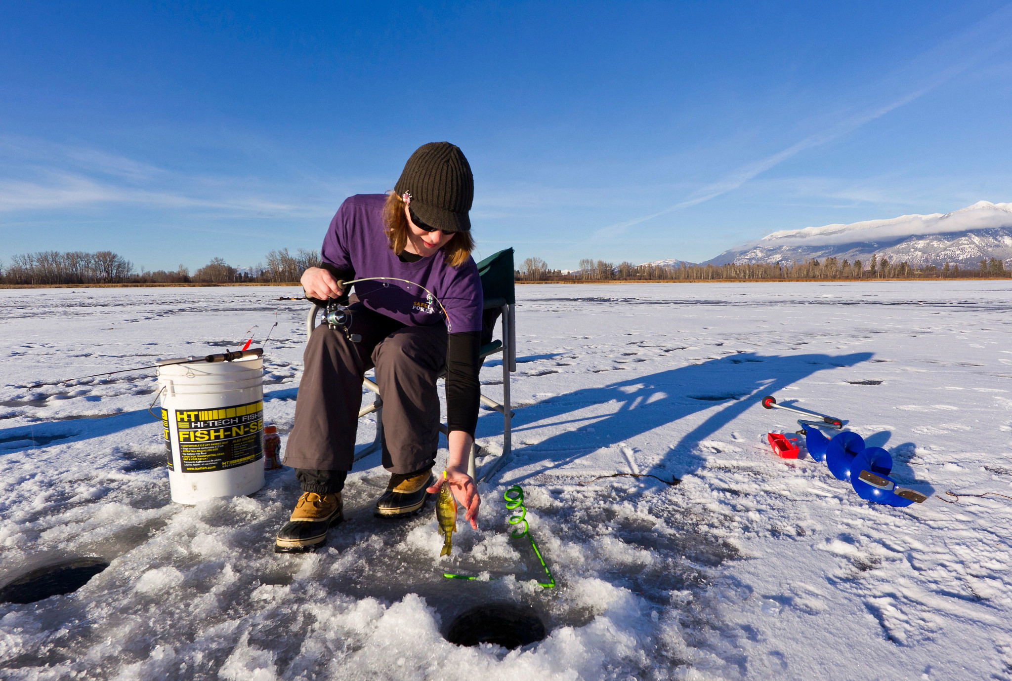 An adult participating in ice fishing on a frozen lake, pulling a fish from an ice hole beside fishing gear and a chair.